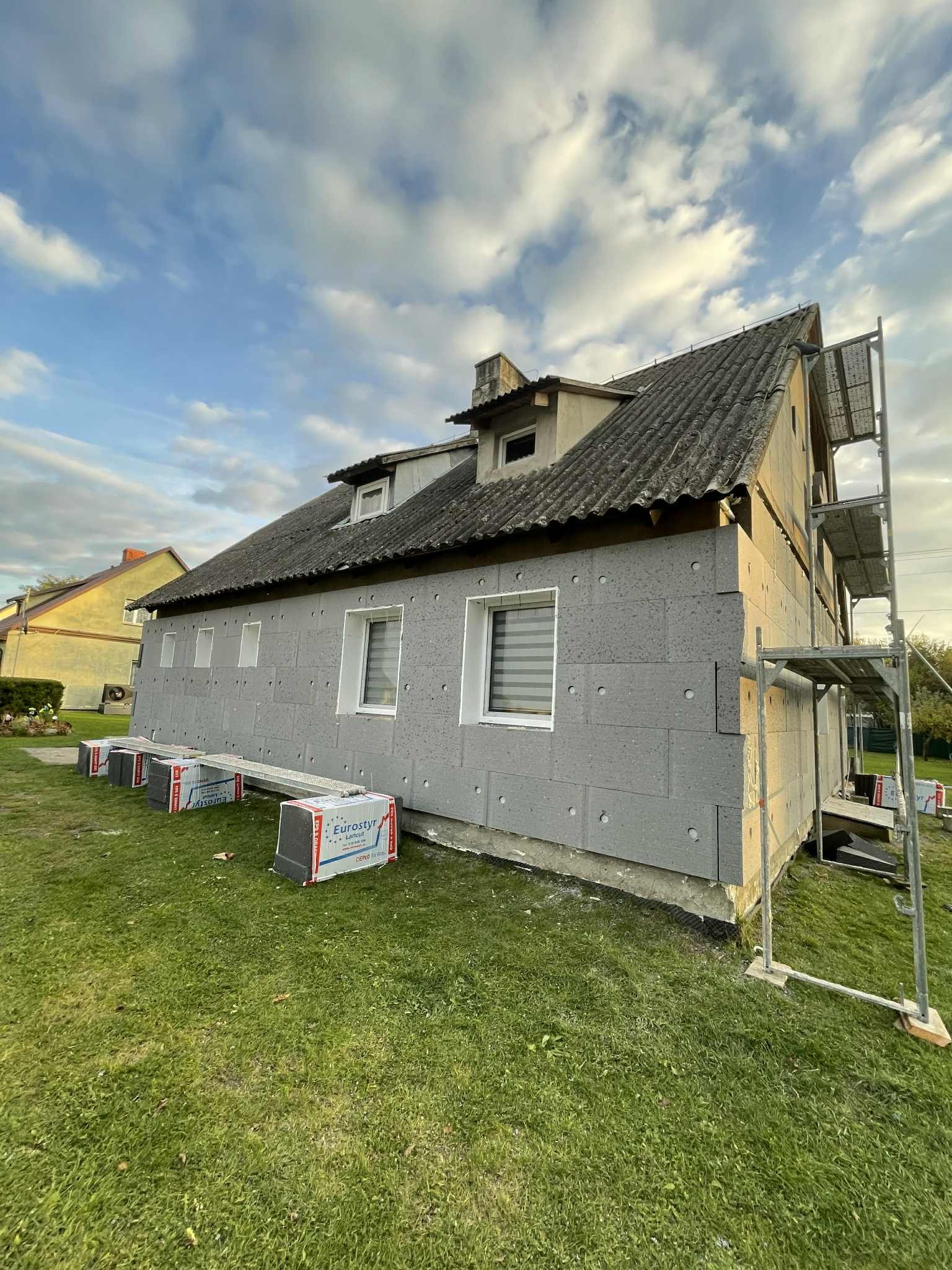 Ocieplenie ścian zewnętrznych domu szarym styropianem, widoczne rusztowanie i paczki styropianu Eurostyr na trawie. Dach pokryty szarą dachówką. Błękitne niebo z chmurami.
