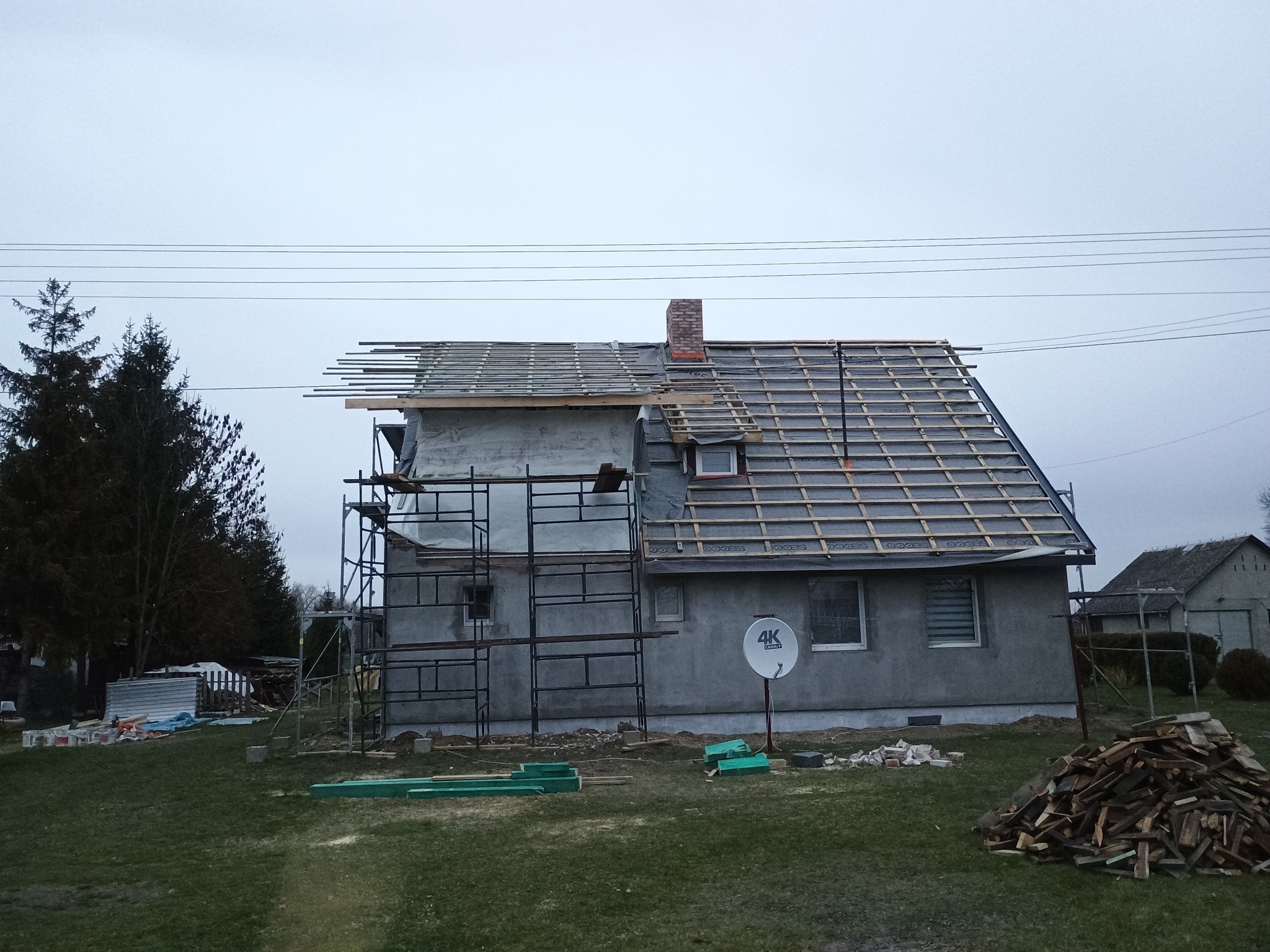 Dom w trakcie krycia dachu, widoczne rusztowania i przygotowane łaty. Część dachu pokryta papą, komin z cegły. Na trawniku leżą drewniane elementy.