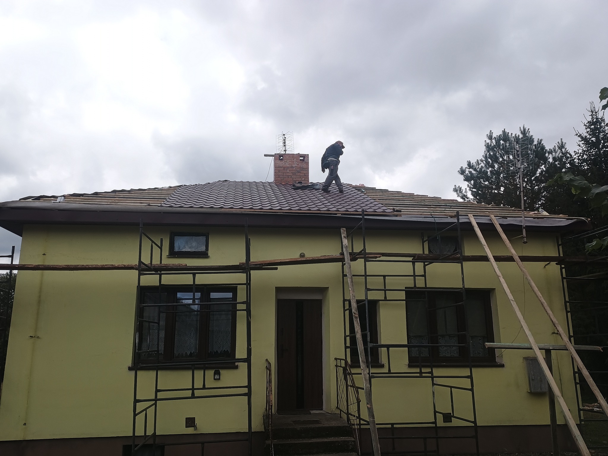 Pracownik na dachu domu jednorodzinnego z brązową dachówką, w trakcie prac dekarskich. Widoczne rusztowania i komin z anteną. Pochmurne niebo w tle.