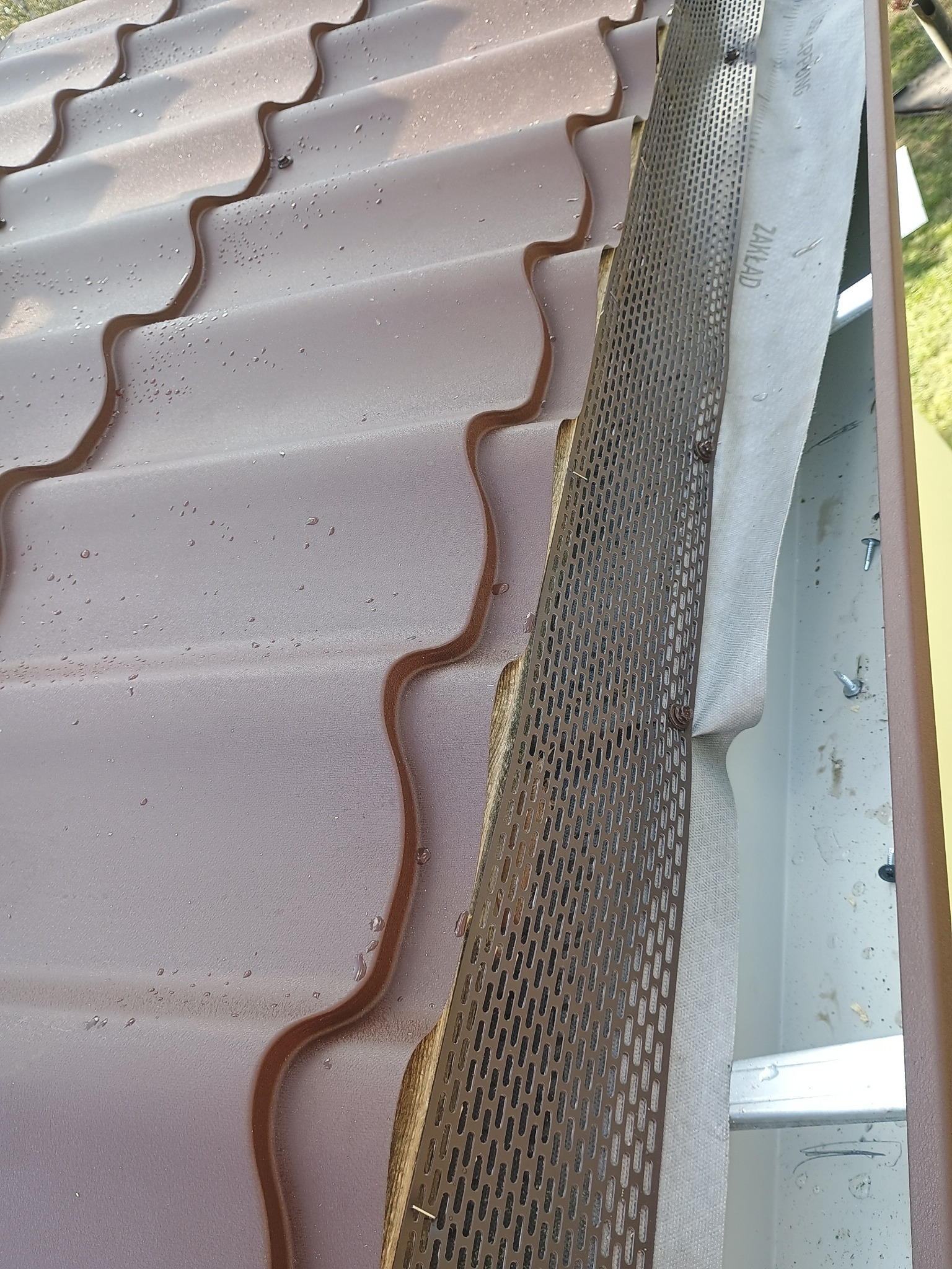 Brązowa blachodachówka z zamontowaną siatką zabezpieczającą rynnę przed liśćmi. Widoczne krople wody na powierzchni dachu.