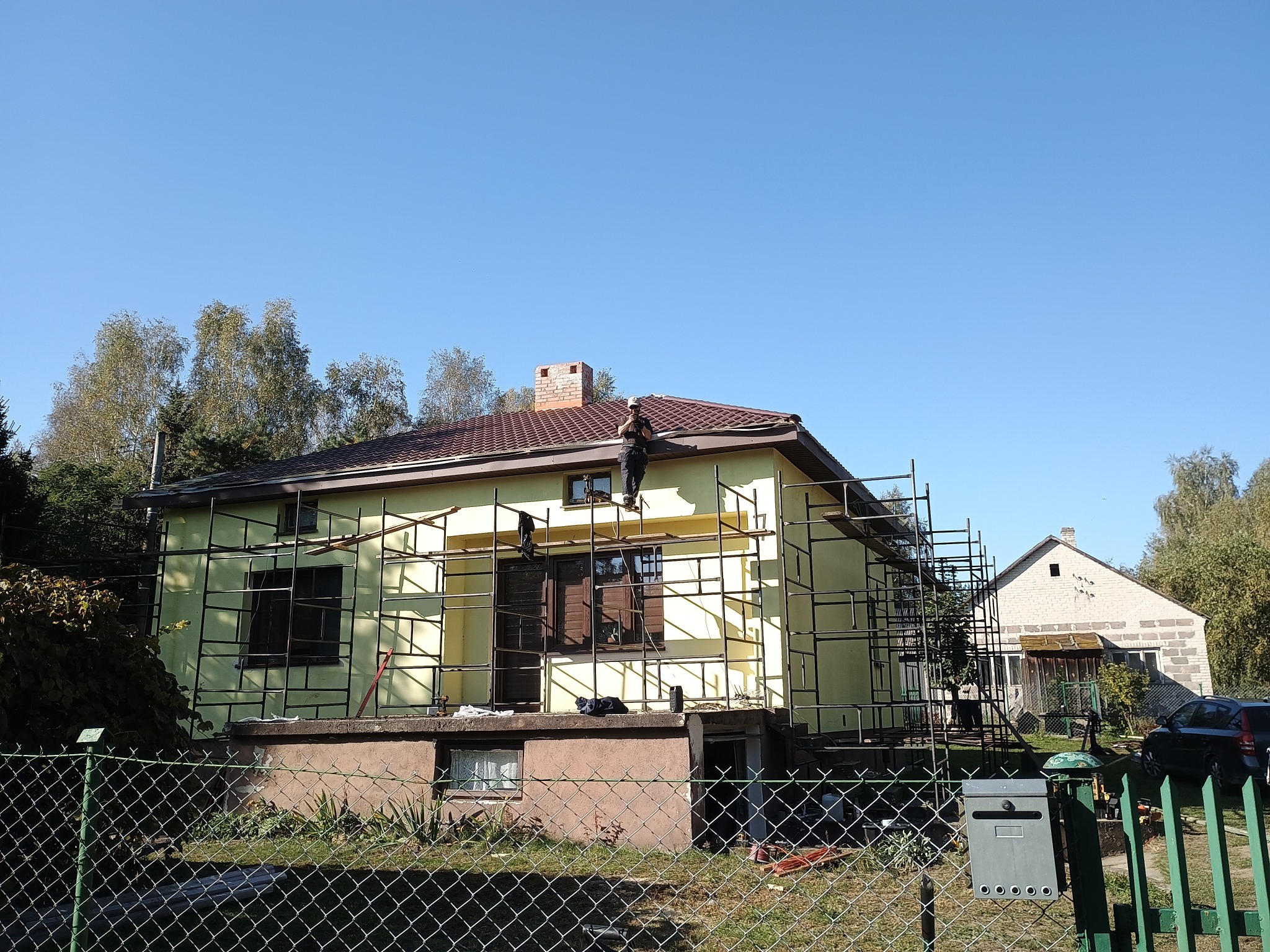 Dom jednorodzinny w trakcie prac dekarskich, z rusztowaniem i pracownikiem na dachu. Widoczny fragment dachu z brązową dachówką i komin z cegły.