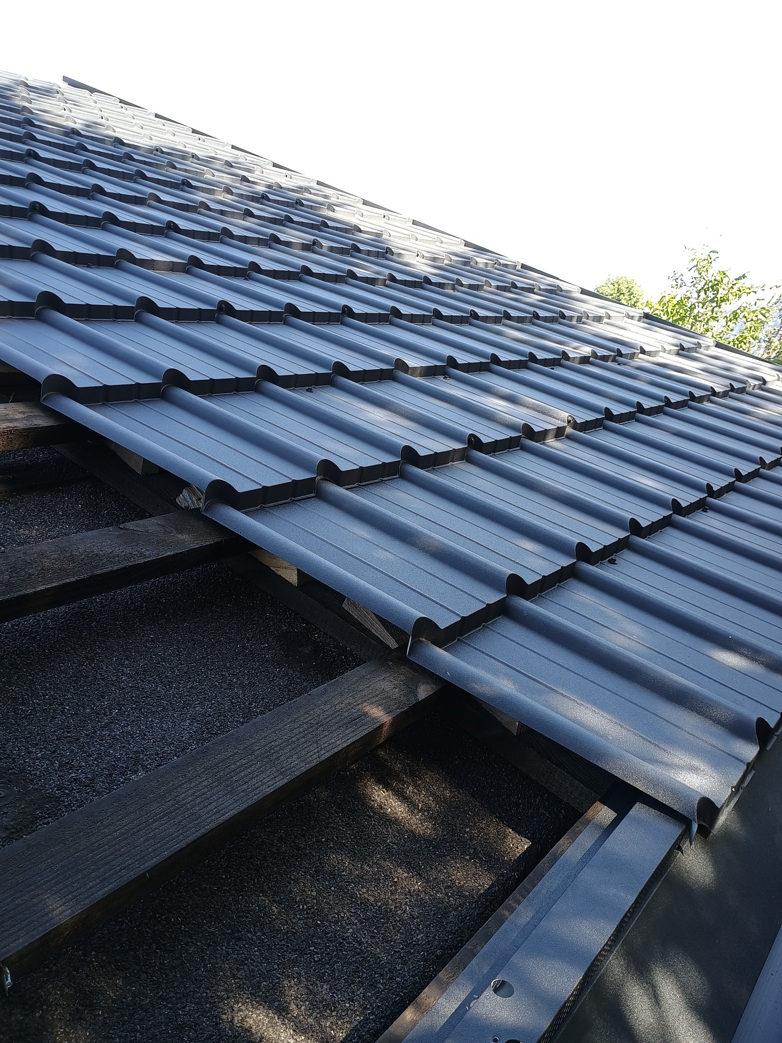 Fragment dachu z szarej blachodachówki trapezowej, widoczne drewniane łaty i podszycie. Ujęcie z dołu pod kątem, w tle zieleń drzew.