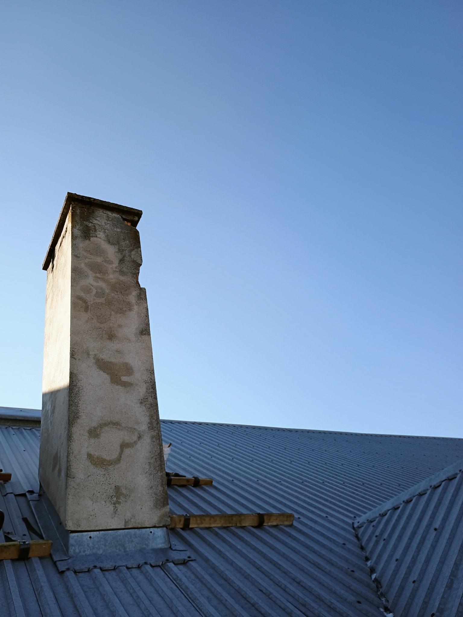 Szary, betonowy komin z ubytkiem na tle blachy trapezowej i błękitnego nieba. Widoczne drewniane elementy konstrukcyjne. Ujęcie z dołu.