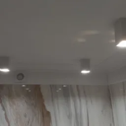 Cztery kwadratowe lampy sufitowe LED, równomiernie rozmieszczone w pomieszczeniu z marmurowymi ścianami i ozdobną sztukaterią. Widoczny wentylator sufitowy.