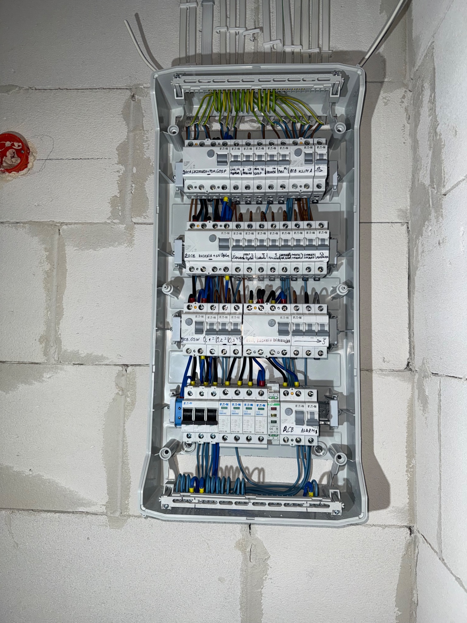 Rozdzielnica elektryczna firmy Eaton z opisami obwodów na ścianie z bloków. Widoczne przewody i bezpieczniki. Instalacja w surowym wnętrzu.