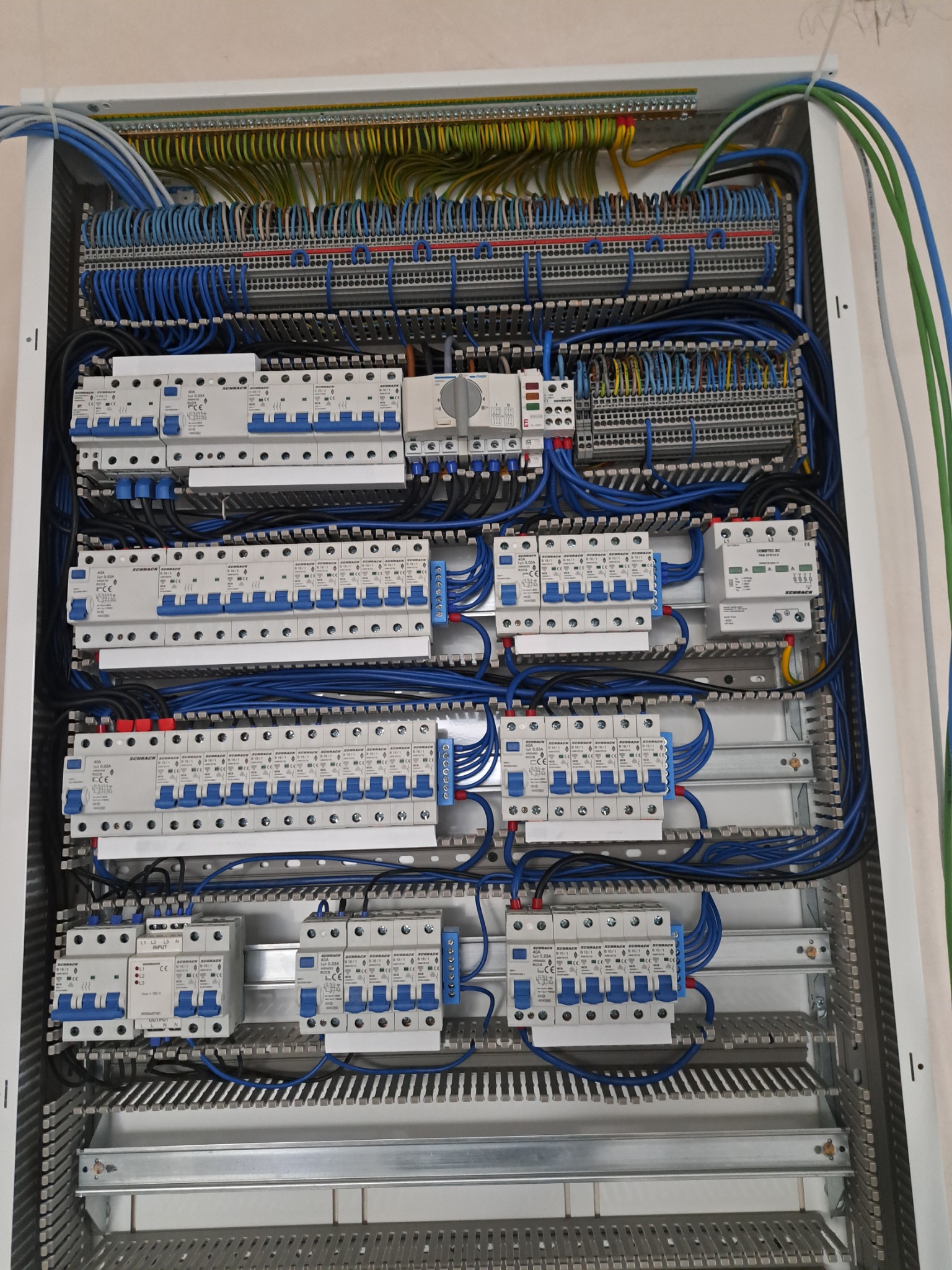 Rozdzielnica elektryczna Schrack z zabezpieczeniami, okablowaniem w kolorach niebieskim, zielonym i żółtym. Widoczne bezpieczniki i listwy zaciskowe.