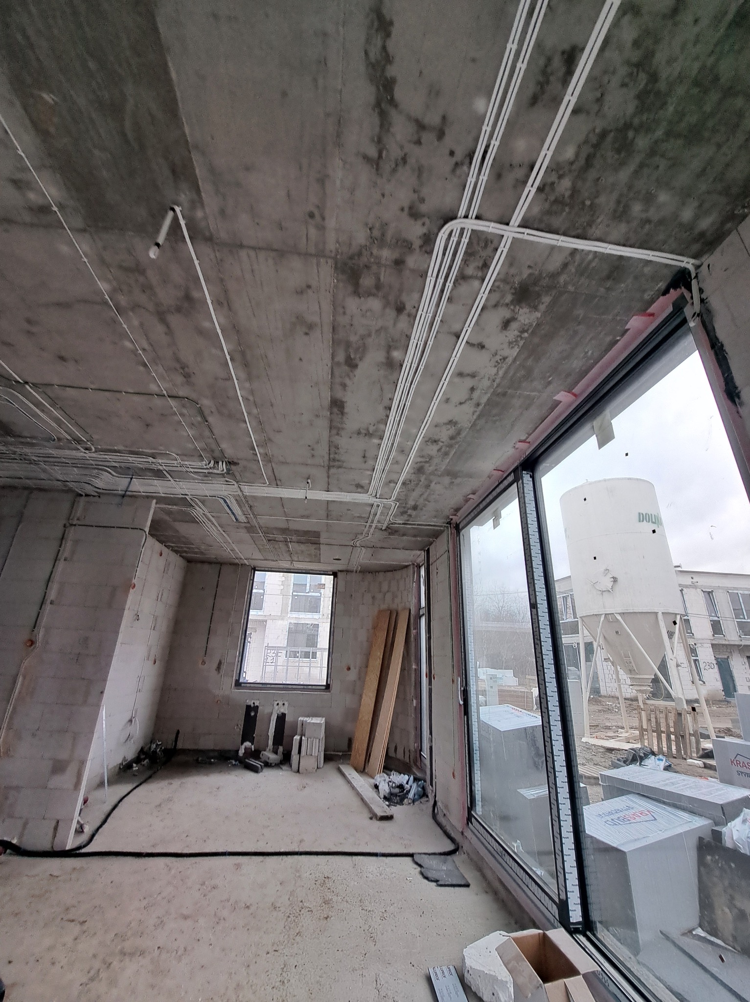 Surowy stan mieszkania z widocznymi instalacjami elektrycznymi na suficie i ścianach. Okno z widokiem na budynek. Białe kable ułożone w peszlach.