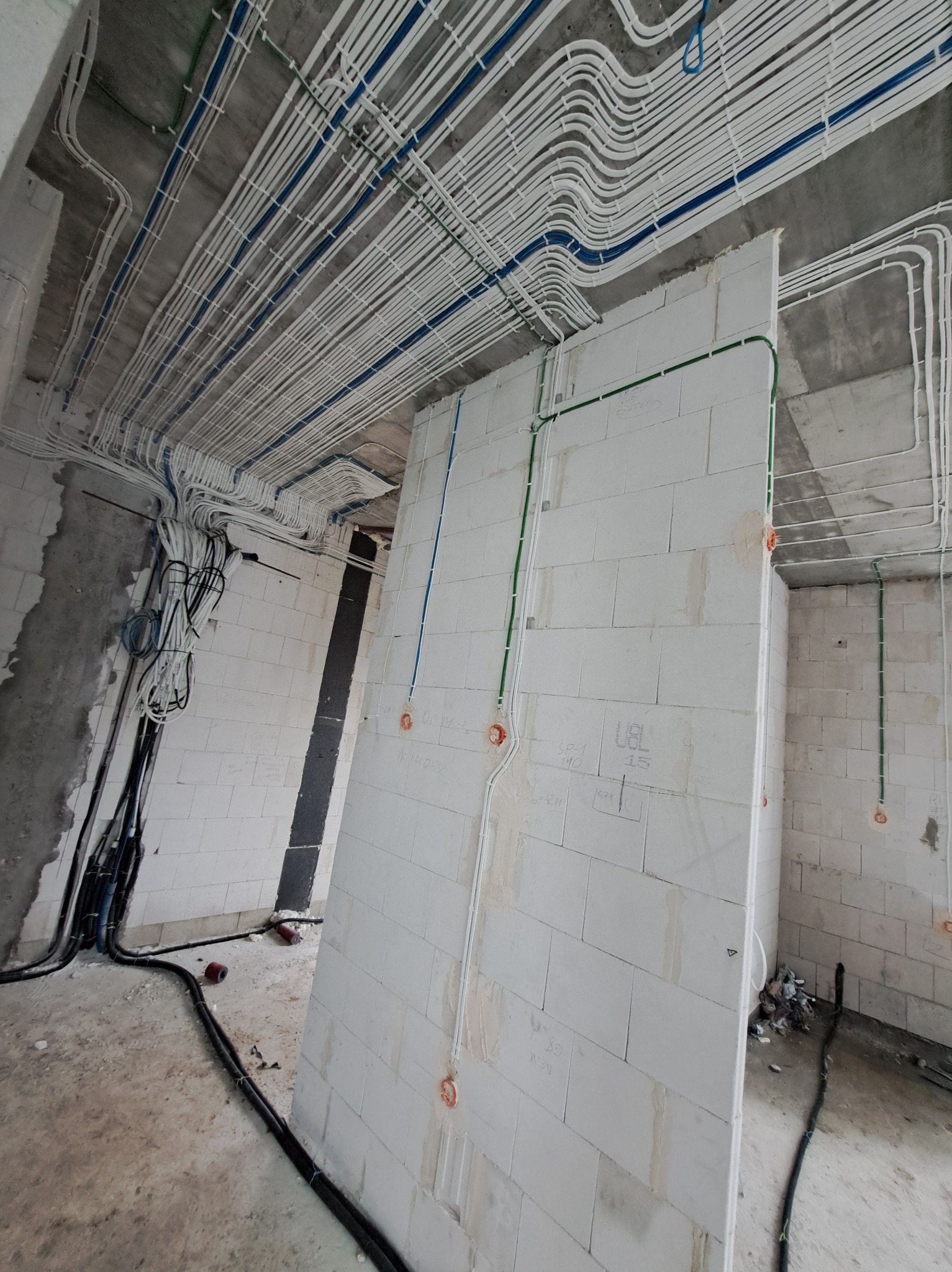 Rozprowadzone kable elektryczne w rurkach na suficie i ścianach budynku w trakcie budowy. Widoczne puszki elektryczne i kable w kolorach: białym, niebieskim i zielonym.