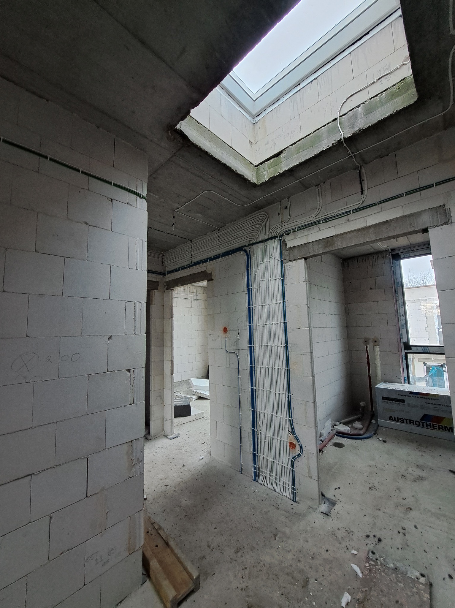 Surowy stan wnętrza z instalacją elektryczną w peszlach, widoczne przewody prowadzone po ścianach z betonu komórkowego, okno dachowe i okno w ścianie.