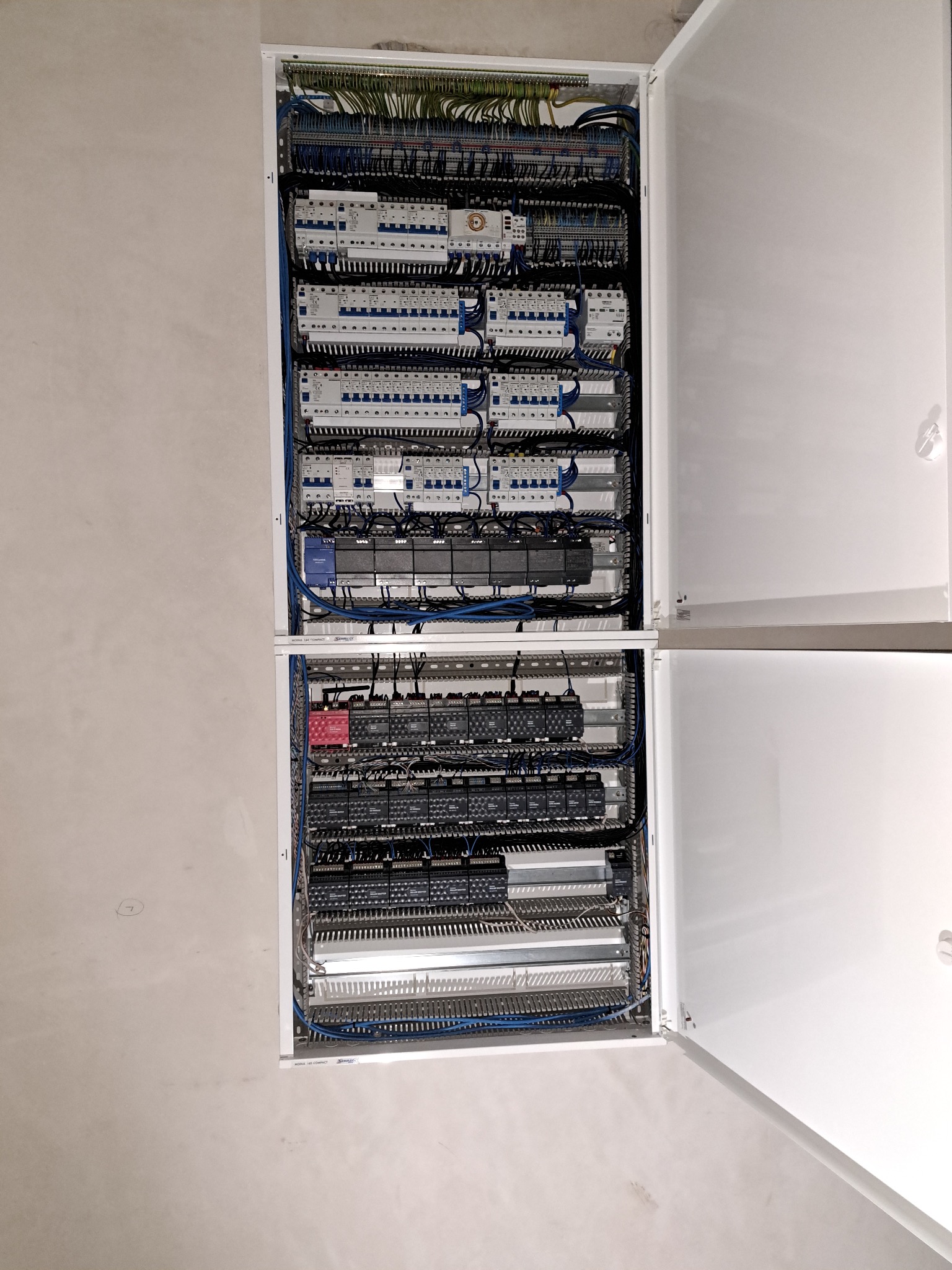 Otwarta szafa elektryczna z licznymi zabezpieczeniami, przewodami i modułami, zamontowana na surowej ścianie. Widoczne detale połączeń i elementów sterujących.