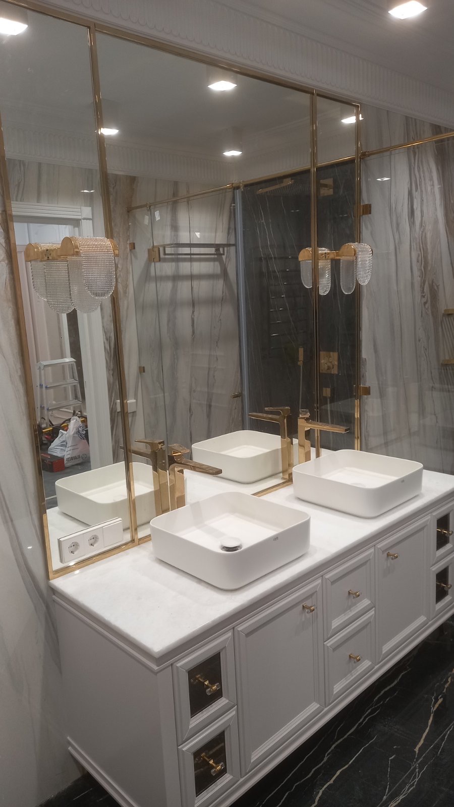 Elegancka łazienka z dwoma białymi umywalkami nablatowymi, złotą armaturą i dużym lustrem w złotej ramie. Widoczne oświetlenie sufitowe i marmurowe wykończenie ścian.