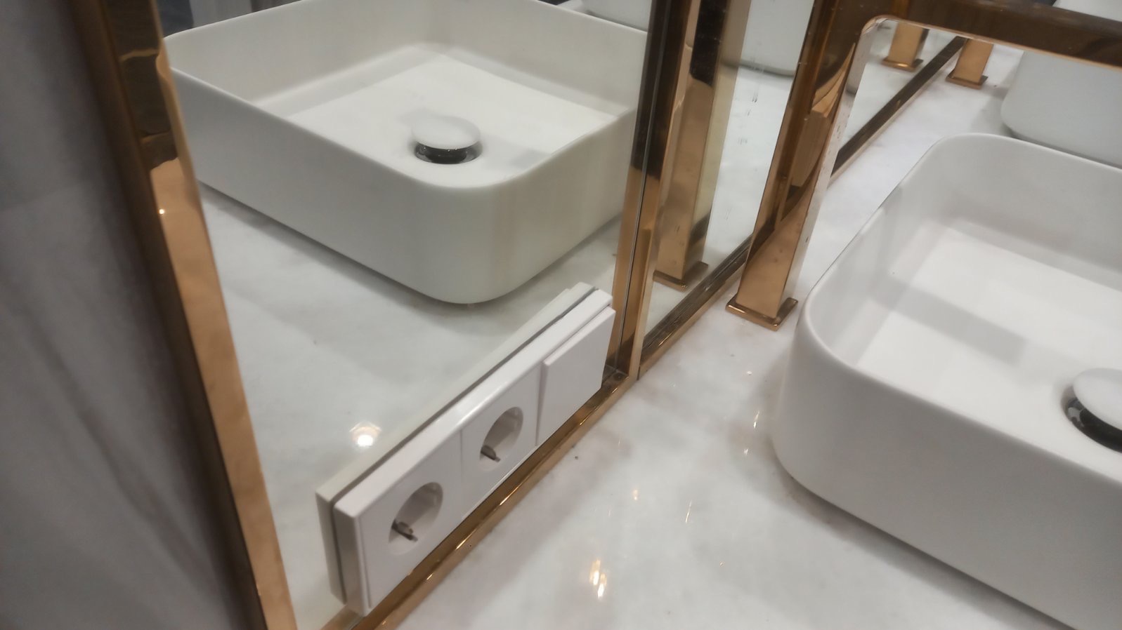 Białe gniazdko elektryczne zamontowane obok umywalki z armaturą w złotym kolorze, odbijające się w lustrze. Widok z bliska, marmurowe wykończenie.