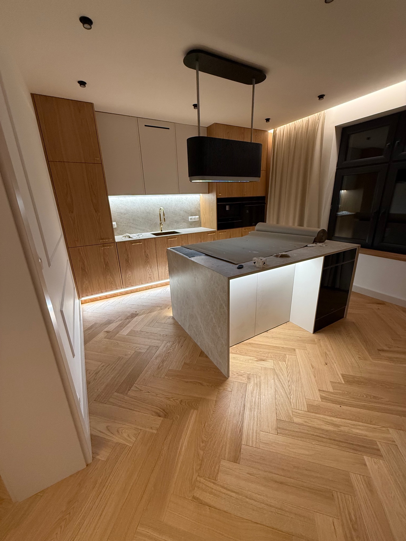 Nowoczesna kuchnia z wyspą i oświetleniem LED pod szafkami. Parkiet ułożony w jodełkę. Na wyspie zabezpieczenie blatu folią. Czarna lampa nad wyspą.