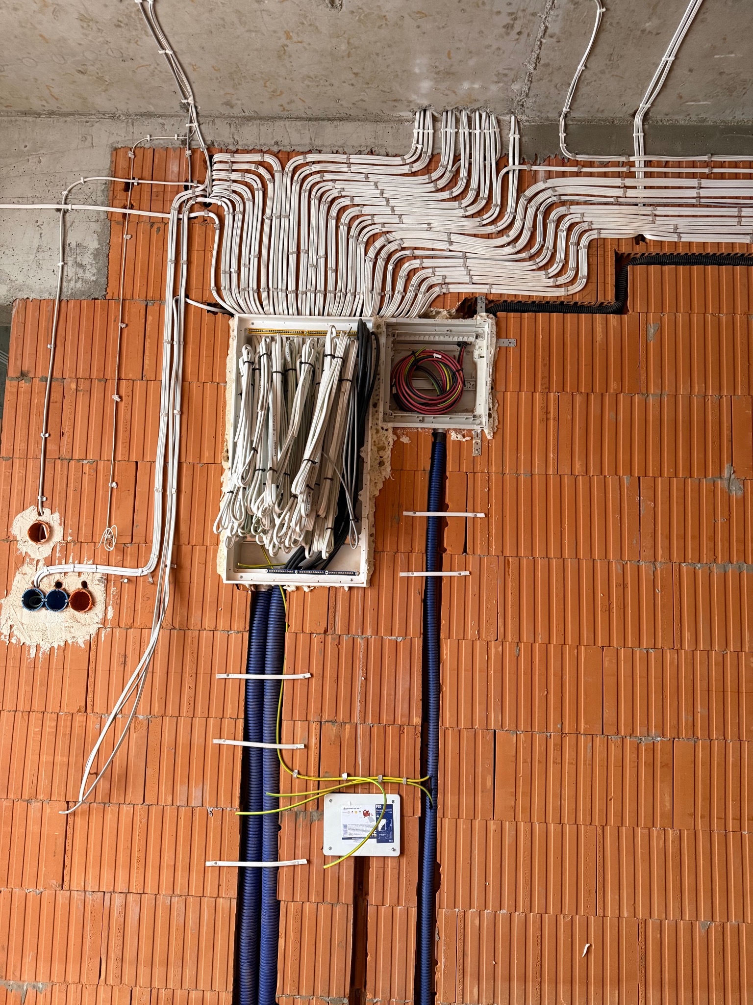 Rozprowadzenie kabli elektrycznych w peszlach i korytkach na ścianie z cegły dziurawki. Widoczne puszki instalacyjne i przewody. Etap prac instalacyjnych.