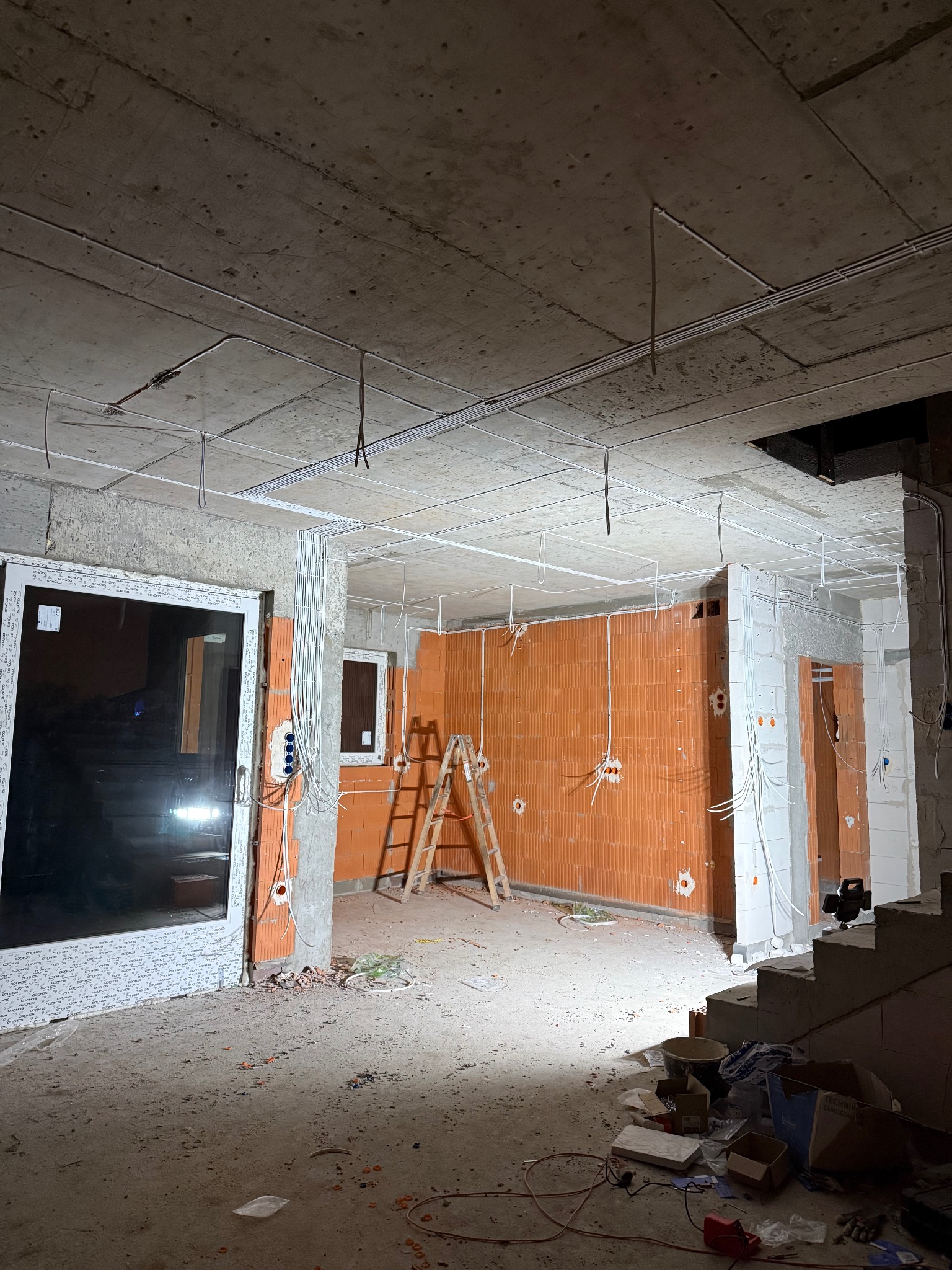 Wnętrze w trakcie prac instalacyjnych: widoczne kable elektryczne prowadzone po suficie i ścianach, ściany z cegły dziurawki, drabina oparta o ścianę, okno i drzwi.