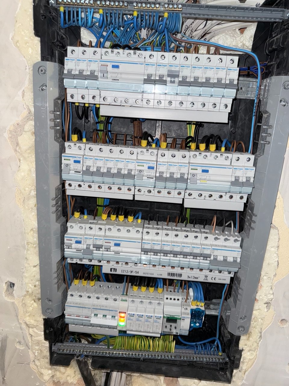 Rozdzielnia elektryczna z zamontowanymi bezpiecznikami i okablowaniem w kolorach: niebieskim, żółto-zielonym i brązowym. Widoczne elementy montażowe i fragment ściany.