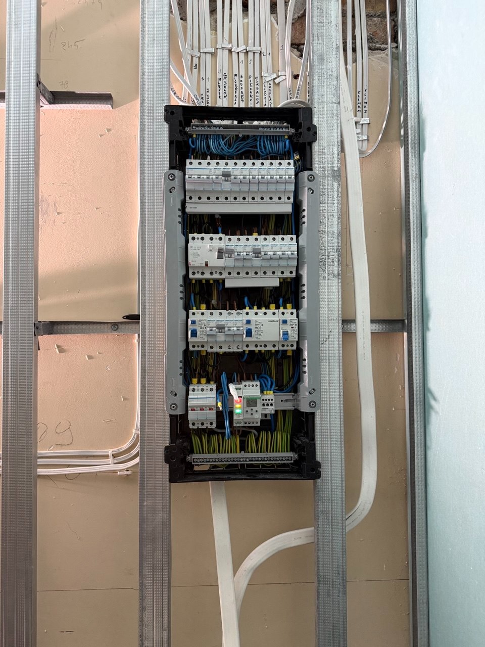 Rozdzielnica elektryczna w trakcie montażu, widoczne kable i zabezpieczenia, zamontowana w metalowej konstrukcji. Częściowo widoczna ściana z instalacją.