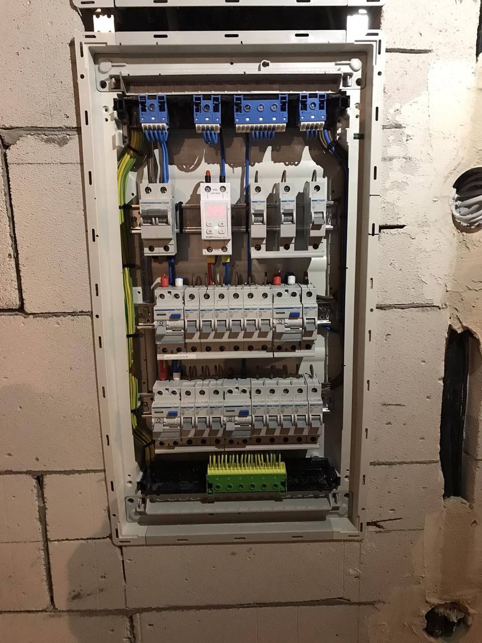 Rozdzielnica elektryczna z zabezpieczeniami, zamontowana na ścianie z betonu komórkowego. Widoczne przewody i elementy instalacji.