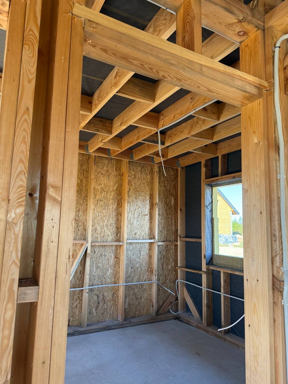 Surowy stan wnętrza domu w budowie: drewniana konstrukcja ścian z płyt OSB, widoczne przewody elektryczne w peszlach, okno z widokiem na zewnątrz.