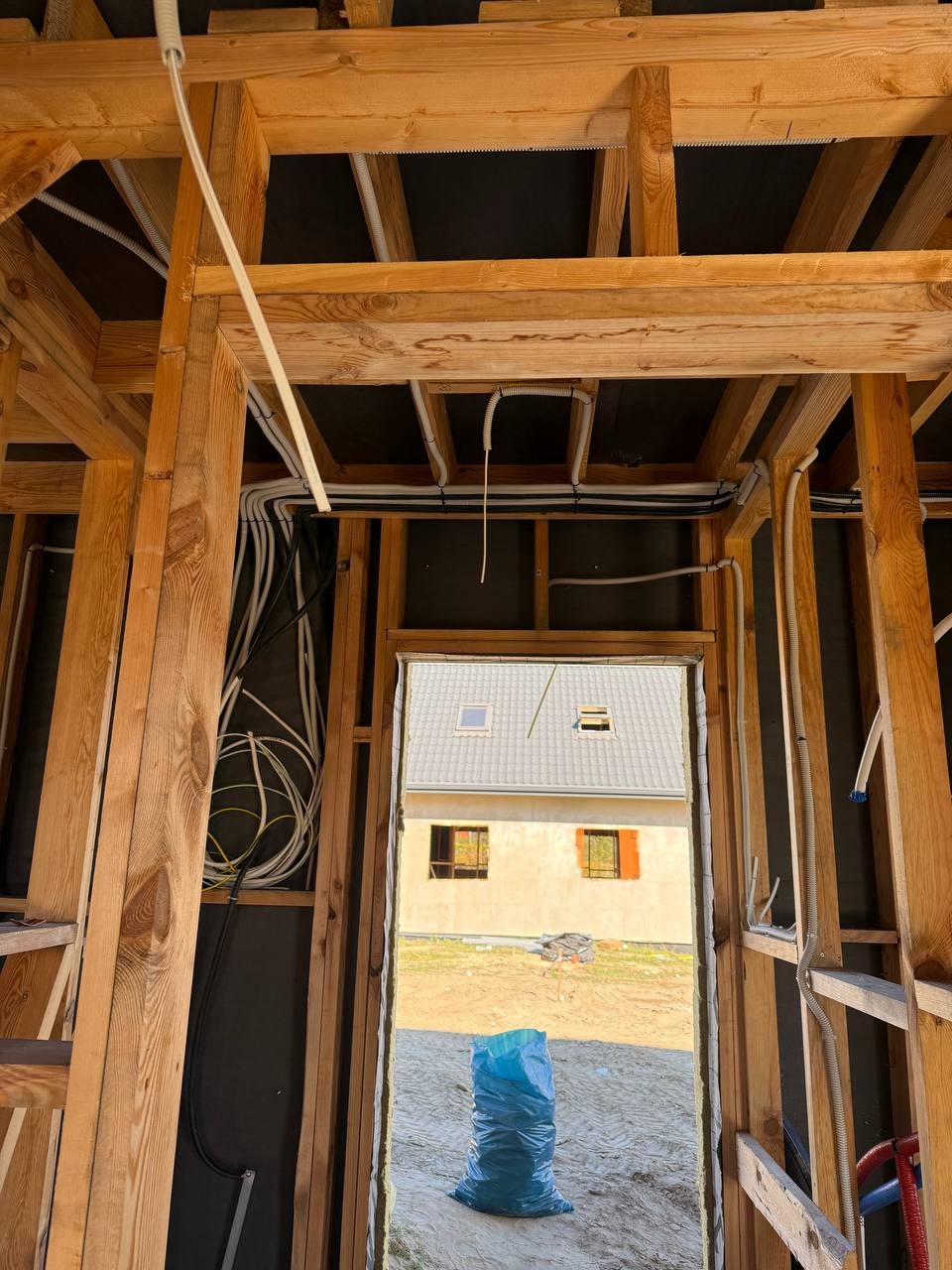 Wnętrze domu w trakcie budowy z widoczną drewnianą konstrukcją i poprowadzonymi przewodami elektrycznymi. W tle widoczny budynek przez otwór drzwiowy.