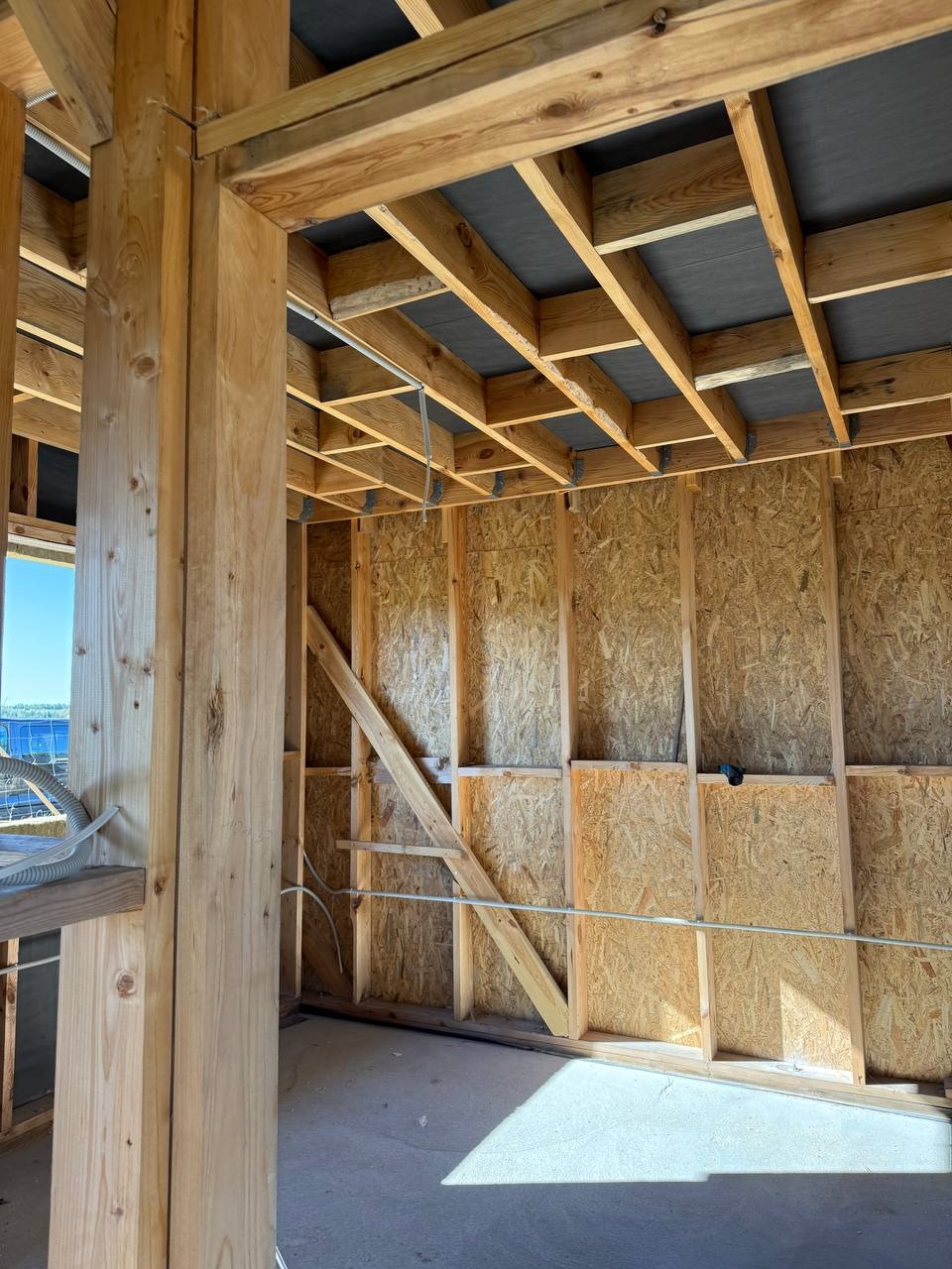 Surowy stan wnętrza domu w budowie z widocznymi drewnianymi belkami konstrukcyjnymi i przewodami elektrycznymi. W tle widoczne okno z niebieskim niebem.