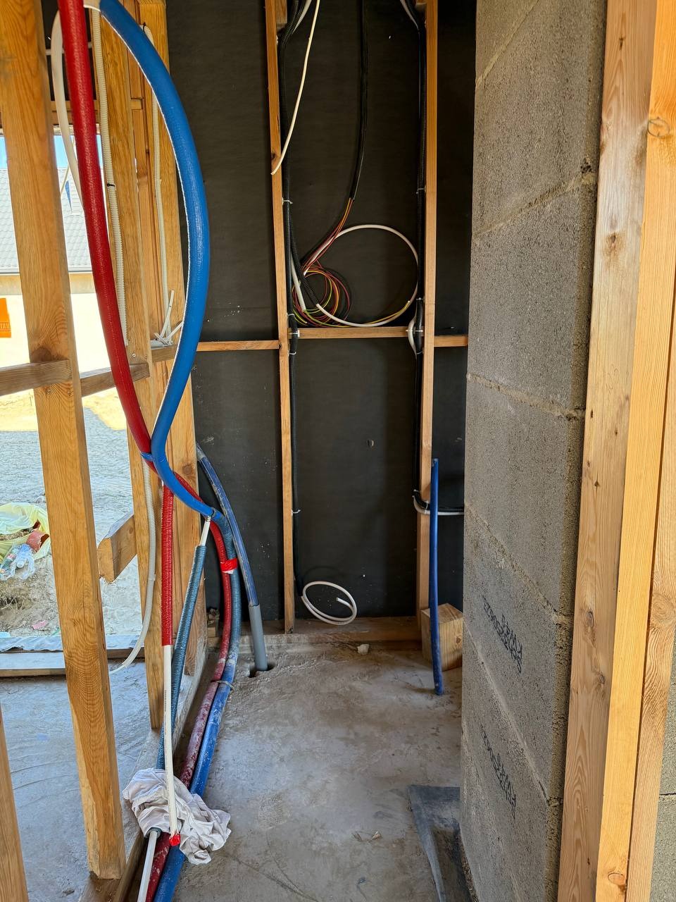 Wnętrze w trakcie budowy: widoczne instalacje wodne (czerwone i niebieskie rury) oraz przewody elektryczne w ścianach z drewnianą konstrukcją i betonowymi bloczkami.