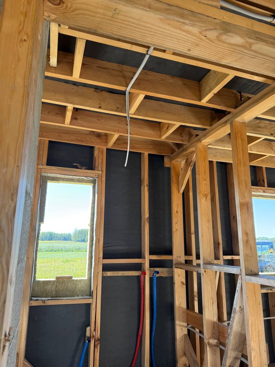 Wnętrze szkieletu domu w budowie z widoczną instalacją elektryczną w peszlu pod sufitem i rurami wodnymi w kolorze niebieskim i czerwonym przy ścianie.