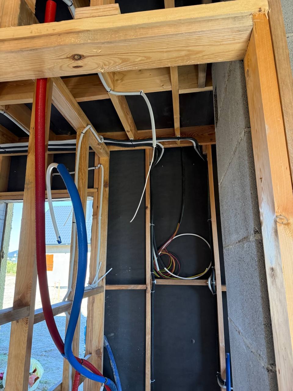 Wnętrze w trakcie budowy z widocznymi instalacjami: rurami w kolorach czerwonym i niebieskim oraz przewodami elektrycznymi w peszlach i bez, zamocowanymi do drewnianej konstrukcji.