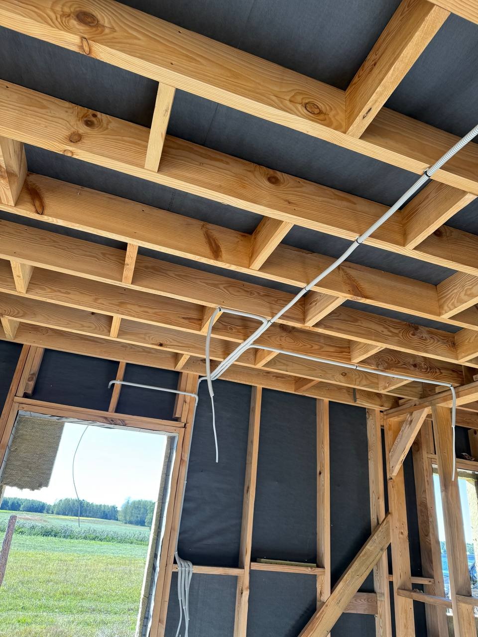 Wnętrze w trakcie budowy z widoczną drewnianą konstrukcją stropu i ścian, oraz poprowadzone kable elektryczne w białych rurkach karbowanych.