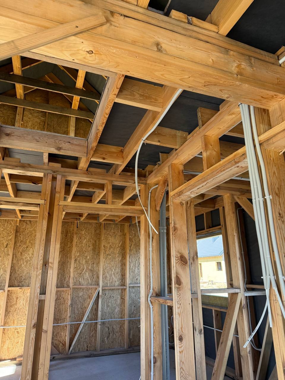 Surowe drewno konstrukcyjne domu w trakcie budowy z widocznymi białymi rurami instalacji elektrycznej. Widok z wnętrza budynku na zewnątrz przez okno.