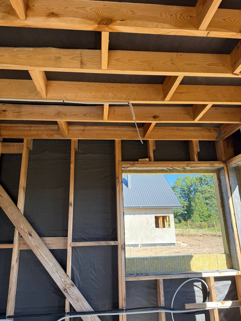 Wnętrze domu w trakcie budowy: drewniana konstrukcja szkieletowa, czarna membrana, ocieplenie, widoczne przewody elektryczne w peszlach. W tle budynek w stanie surowym.