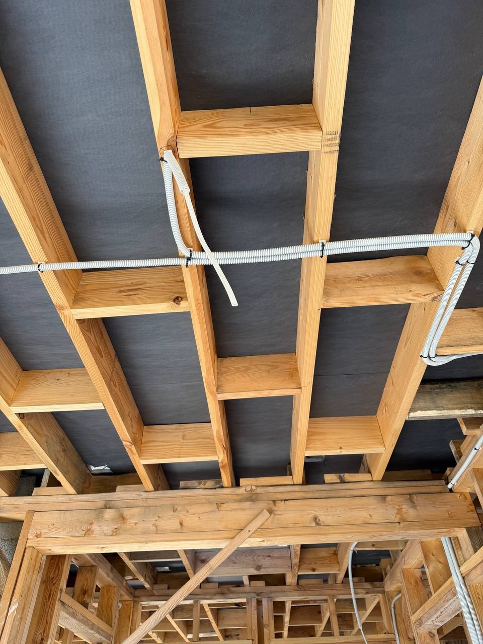 Białe rury w karbowanej osłonie przymocowane do drewnianej konstrukcji stropu z czarną membraną, widok z dołu, etap prac instalacyjnych.