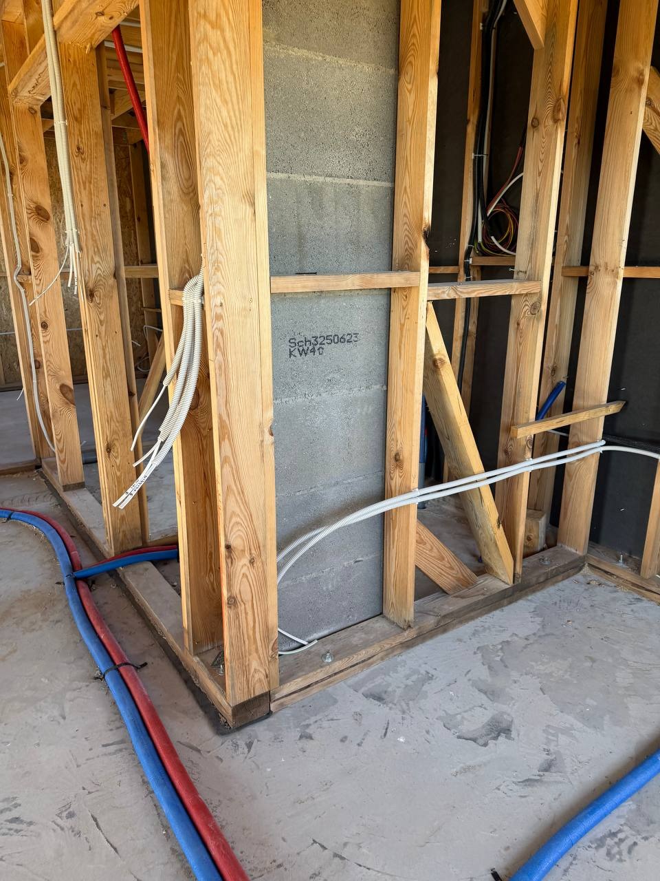Surowy stan: drewniana konstrukcja ściany z instalacjami (rury, kable) wewnątrz. Widoczne szare bloczki betonowe i fragmenty podłogi. Wnętrze w trakcie budowy.