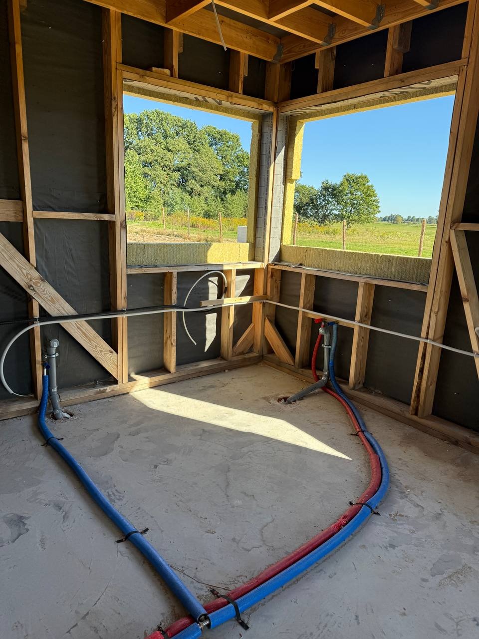 Wnętrze w stanie surowym z widoczną instalacją wodną (rury niebieskie i czerwone) ułożoną na posadzce. W tle okno z widokiem na zieleń.