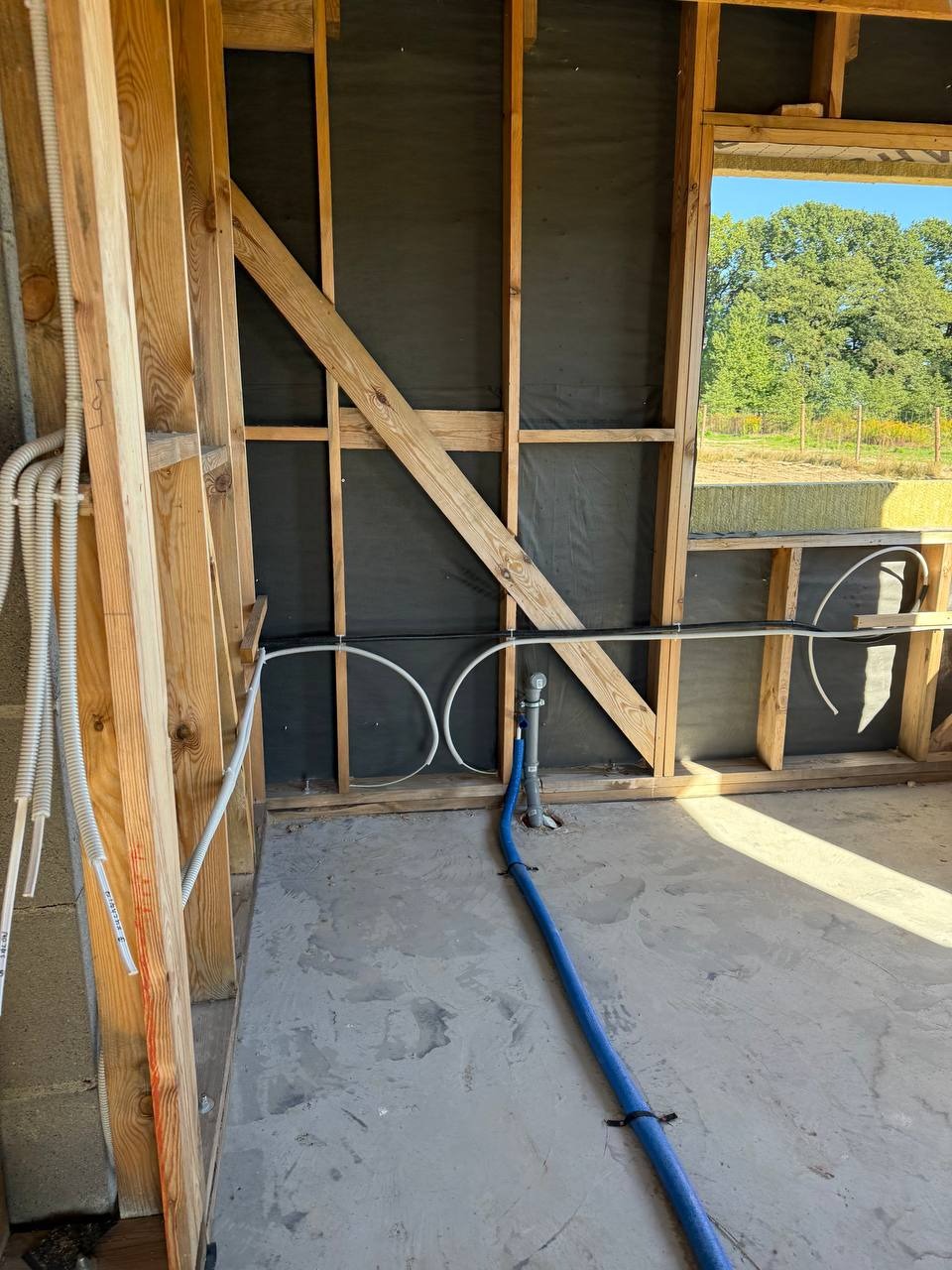 Surowy stan: drewniany szkielet budynku z widocznymi przewodami elektrycznymi i rurami, czarna membrana, betonowa podłoga. Widok na zieleń przez okno.