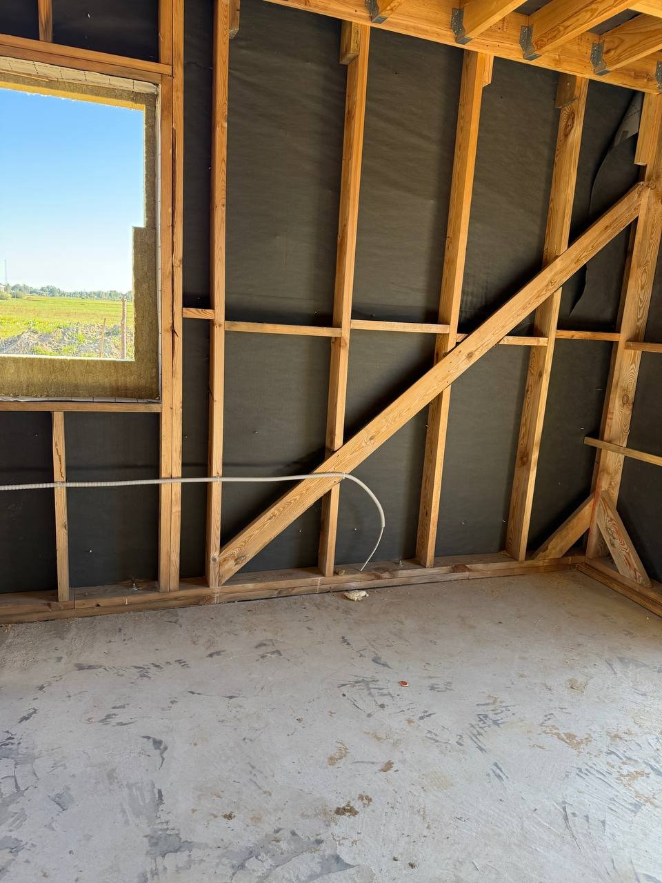Wnętrze w trakcie budowy: drewniana konstrukcja ścian z czarną membraną, widoczne przewody elektryczne, surowy beton na podłodze, okno z widokiem na zewnątrz.
