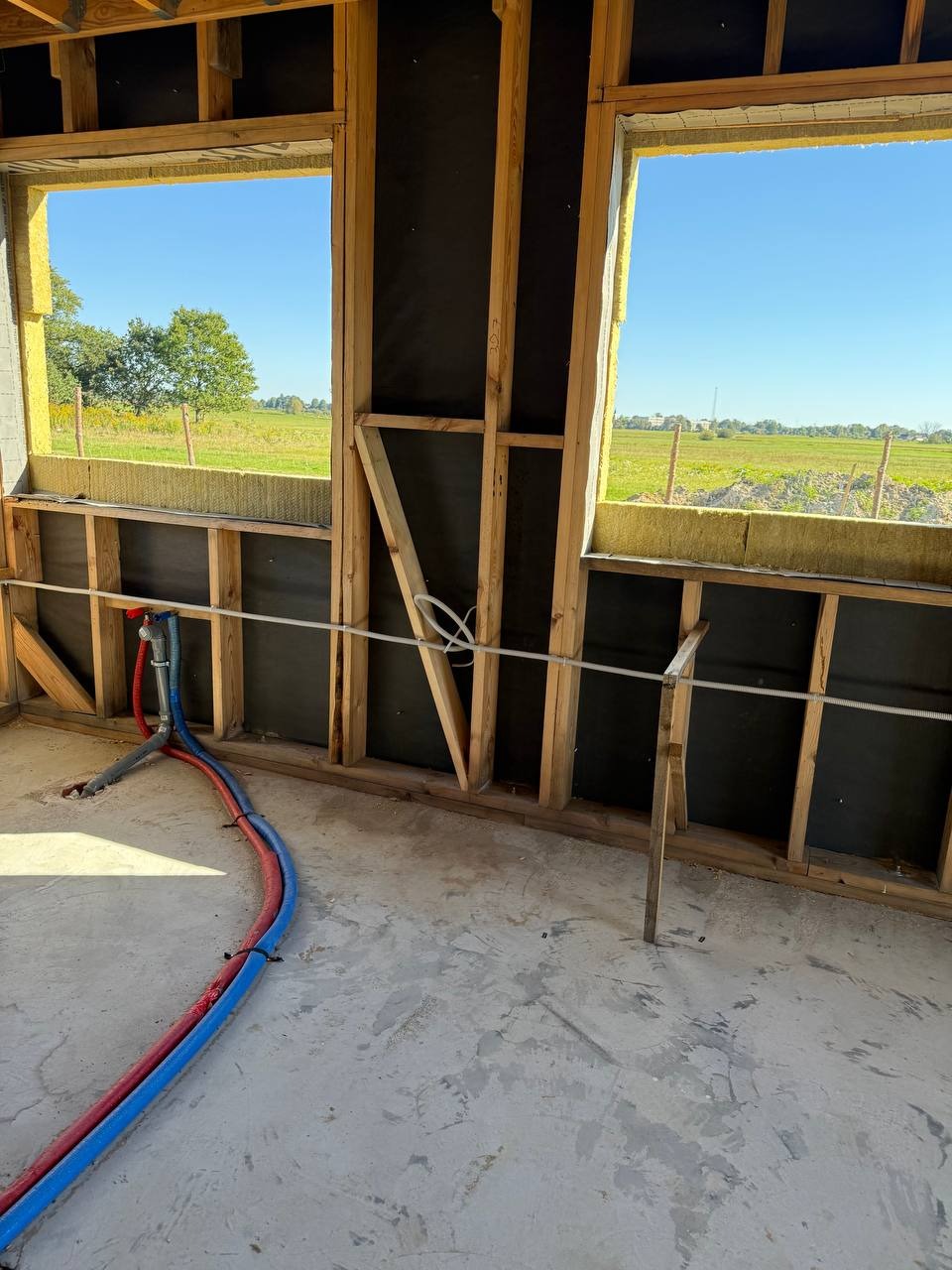 Wnętrze w trakcie budowy: widoczne drewniane ramy okienne z izolacją, czarna folia, betonowa podłoga i rury instalacyjne w kolorach niebieskim i czerwonym.