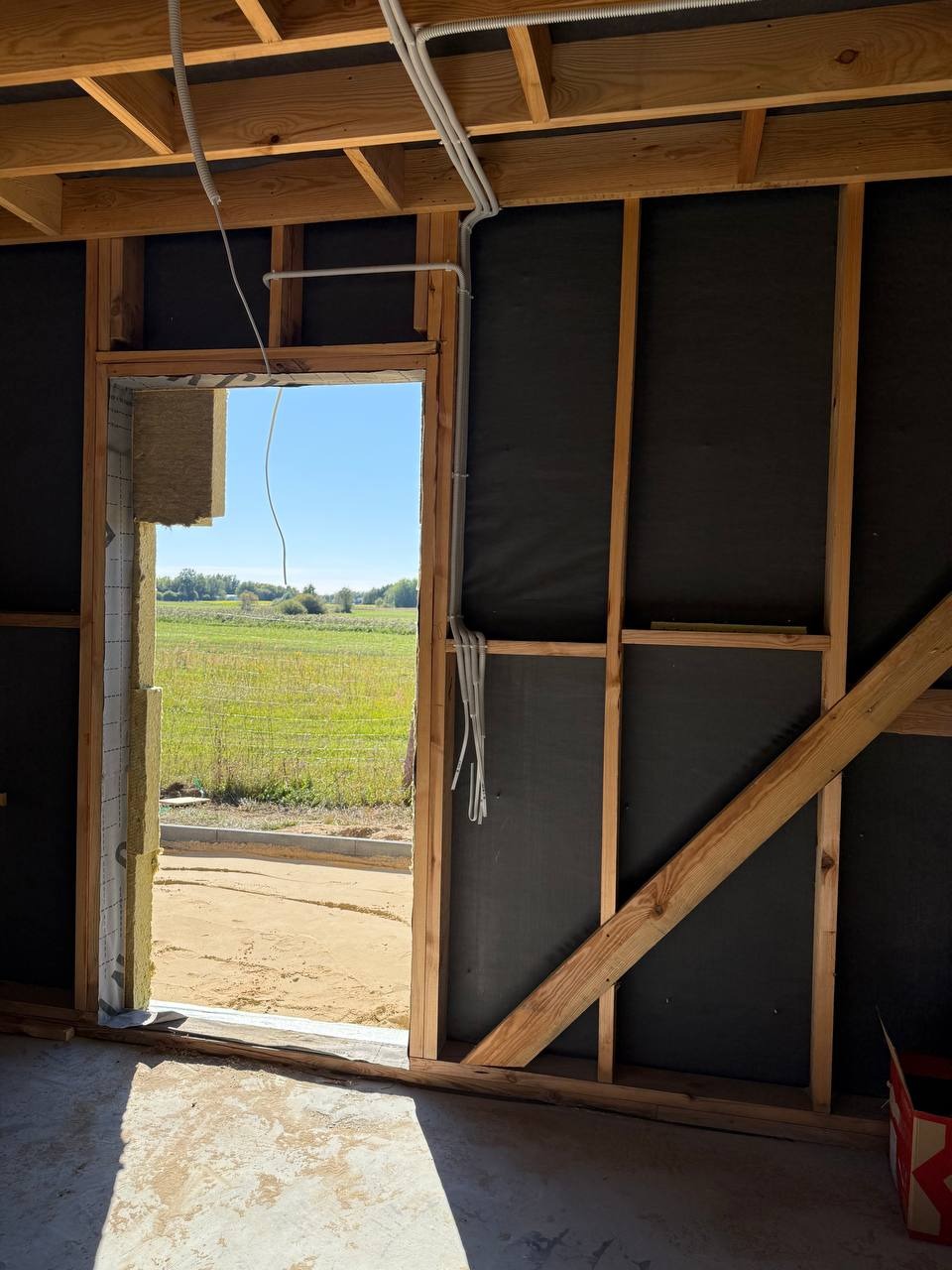 Surowe wnętrze w budowie w Warszawie: widoczne kable elektryczne prowadzone po drewnianej konstrukcji ściany, okno z widokiem na zewnątrz, izolacja.