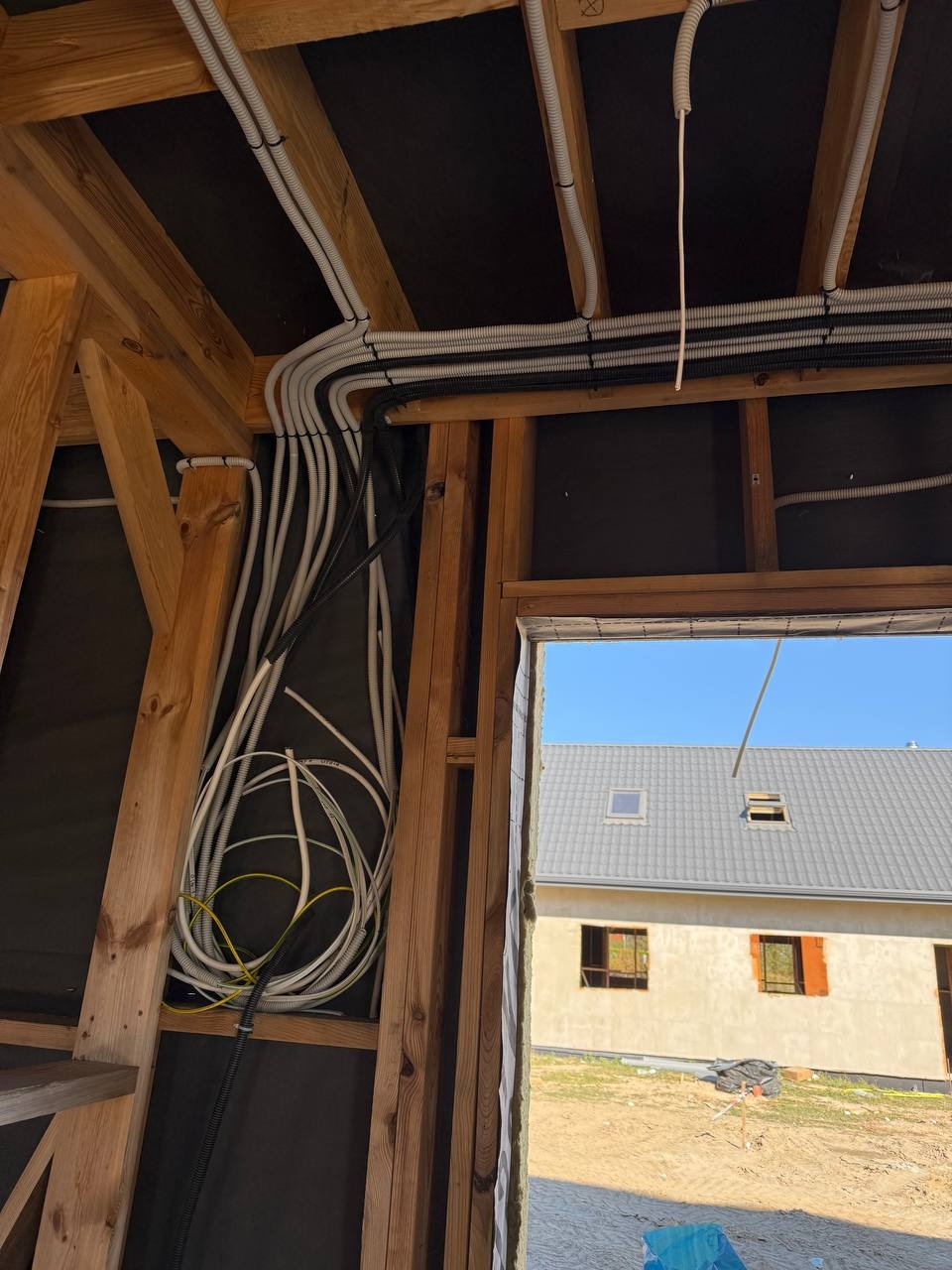 Wnętrze w trakcie budowy z widocznymi kablami elektrycznymi ułożonymi wzdłuż drewnianej konstrukcji. W tle widoczny budynek przez otwór okienny.