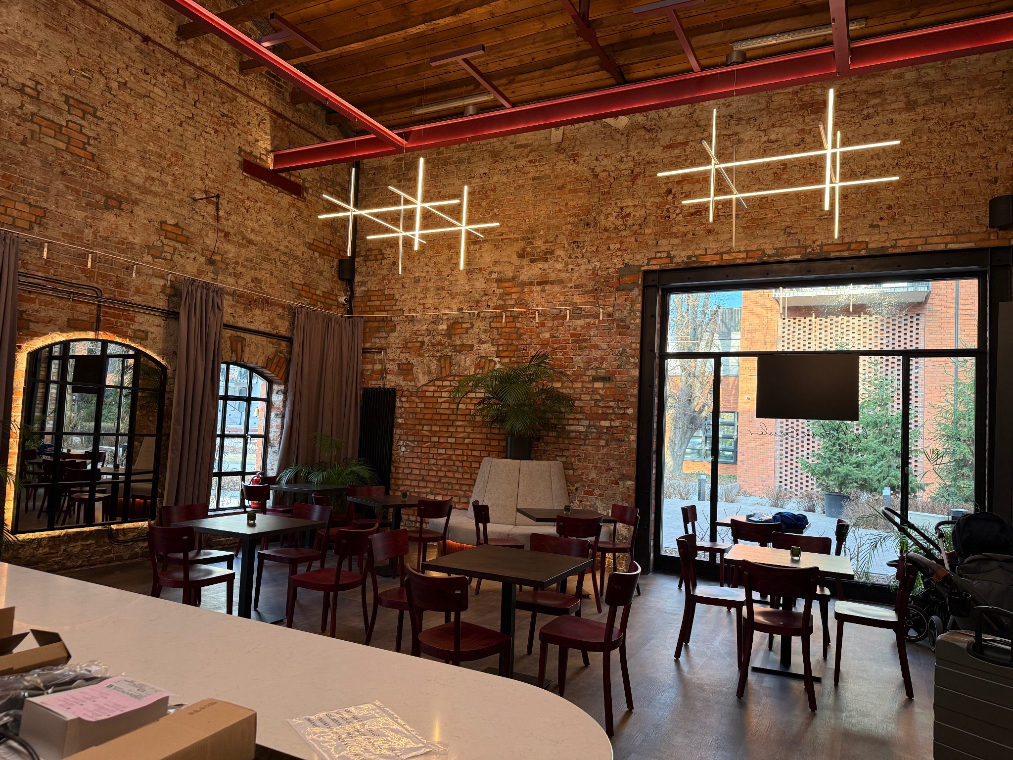 Wnętrze restauracji z ceglanymi ścianami i nowoczesnym oświetleniem w kształcie krzyży. Widoczne stoliki z krzesłami i duże okna z widokiem na zewnątrz.