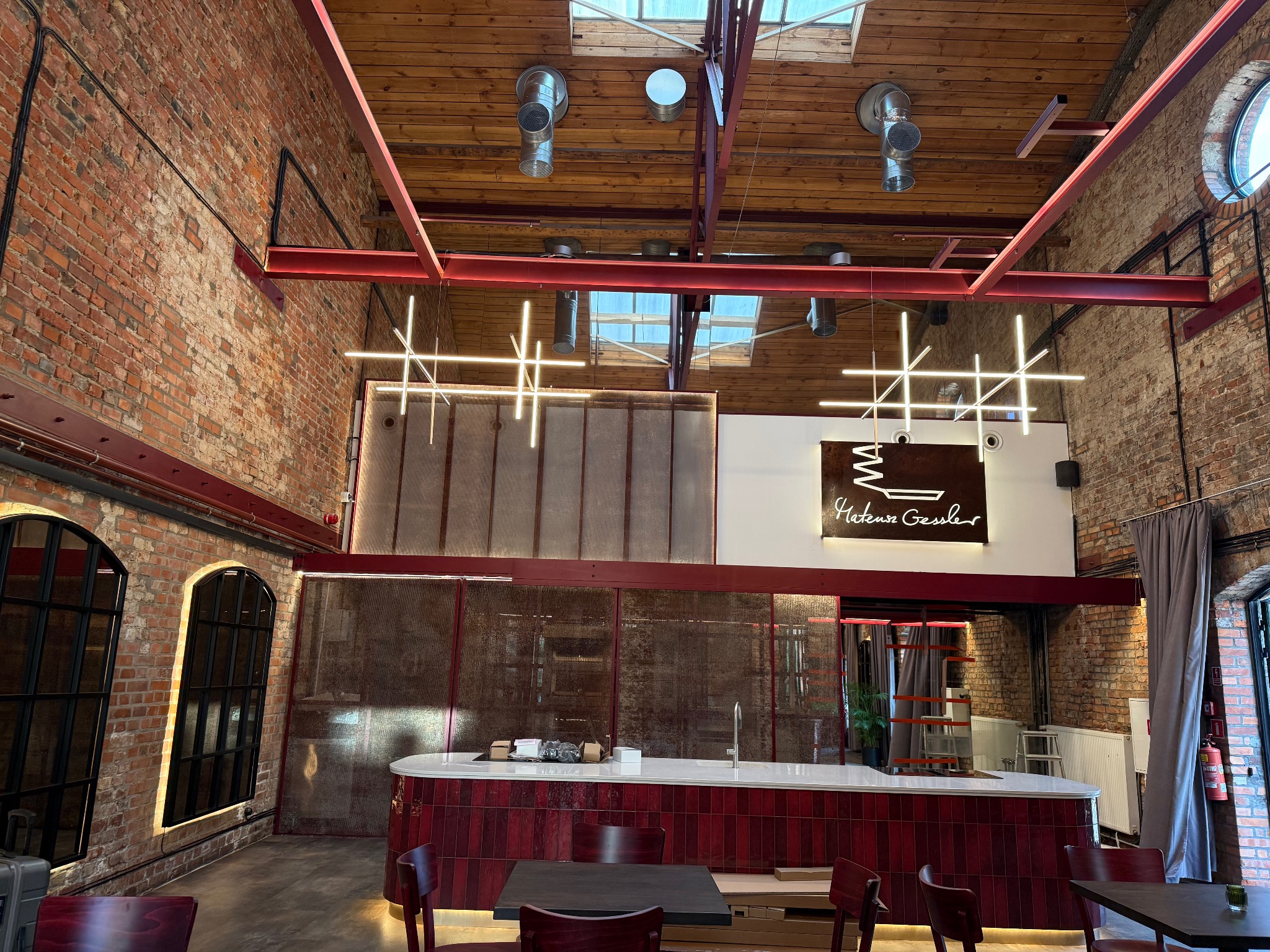 Loftowe wnętrze restauracji z ceglanymi ścianami i nowoczesnym oświetleniem w postaci geometrycznych lamp wiszących. Widoczne elementy instalacji pod sufitem.