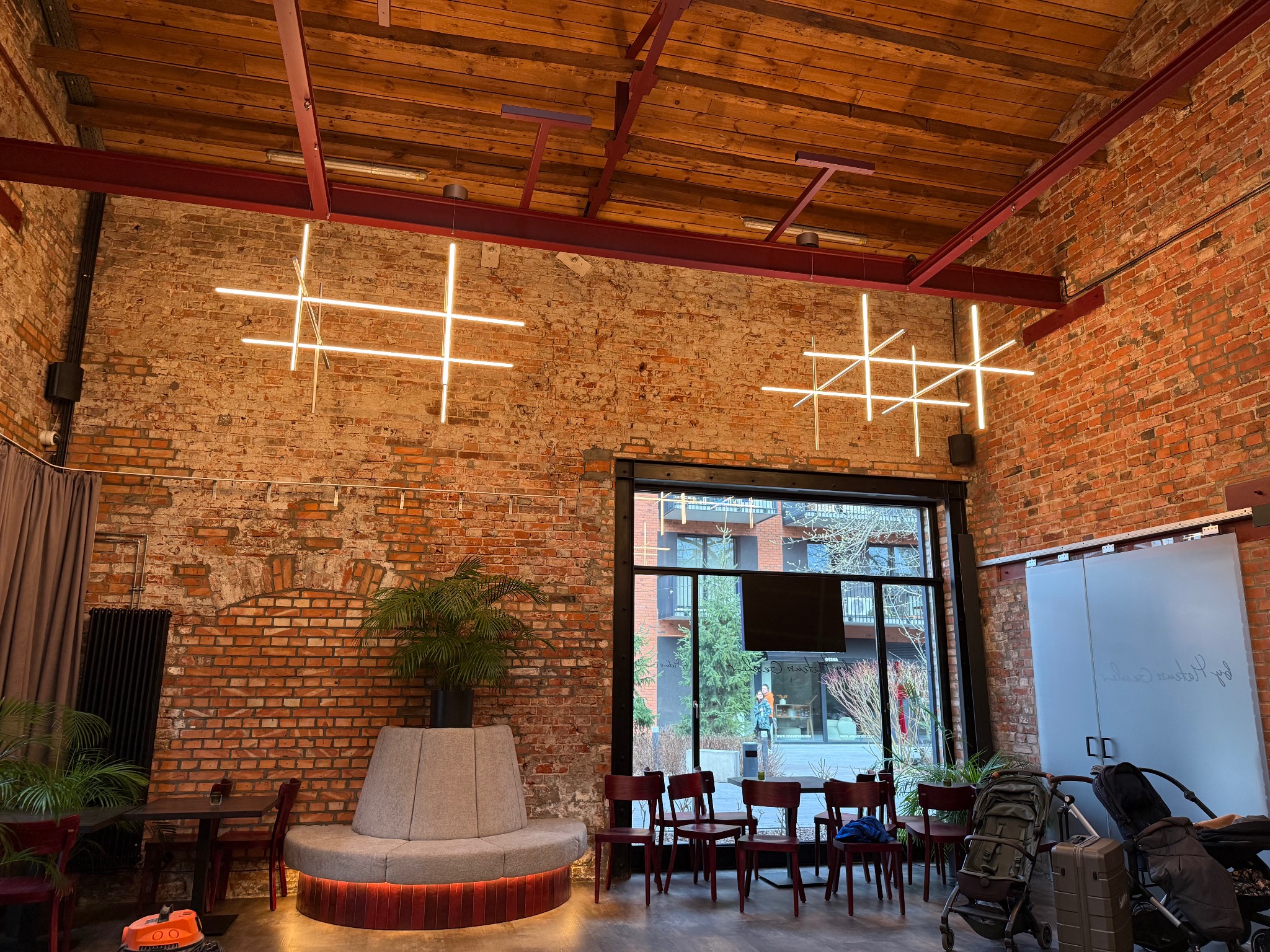 Loftowe wnętrze z ceglaną ścianą i geometrycznymi lampami krzyżowymi. Widoczne drewniane belki stropowe i duże okno z widokiem na zewnątrz. Nowoczesny design oświetlenia.