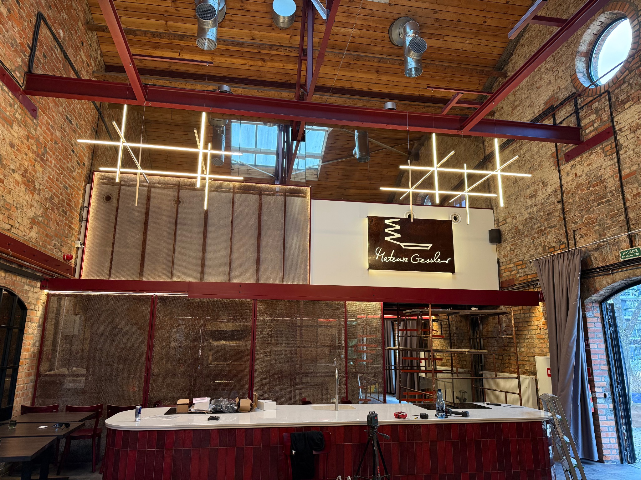 Nowoczesne oświetlenie LED w loftowym wnętrzu restauracji z ceglanymi ścianami i drewnianym stropem. Geometryczne lampy krzyżowe nad barem.