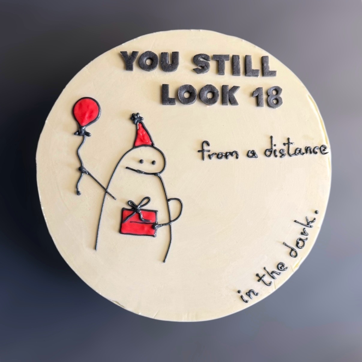 Okrągły tort z kremową polewą, dekorowany zabawnym rysunkiem i napisem 'You still look 18 from a distance in the dark'.