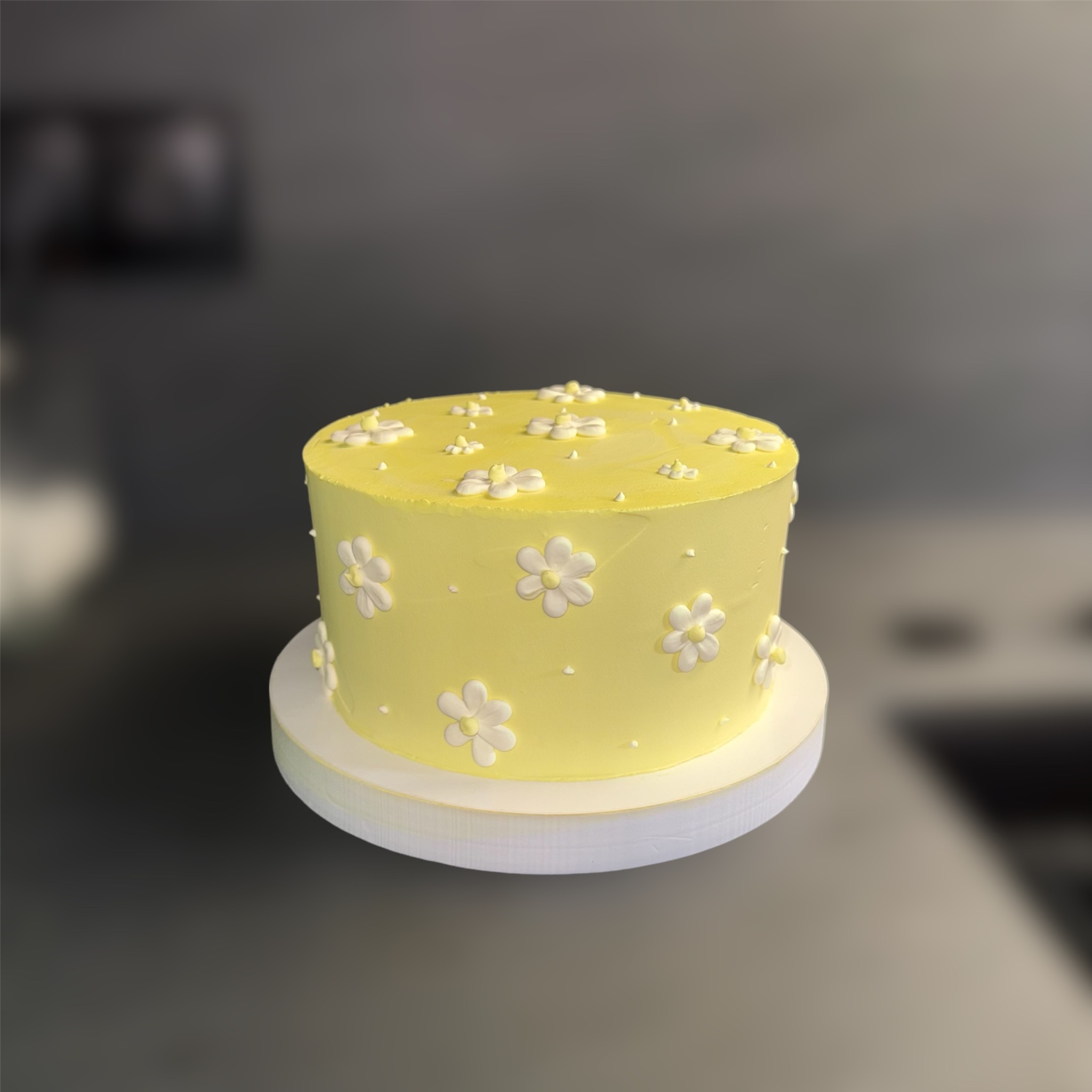 Żółty tort z kremem, ozdobiony białymi kwiatkami na białym podkładzie. Gładka powierzchnia i delikatne zdobienia tworzą elegancki, minimalistyczny wygląd.