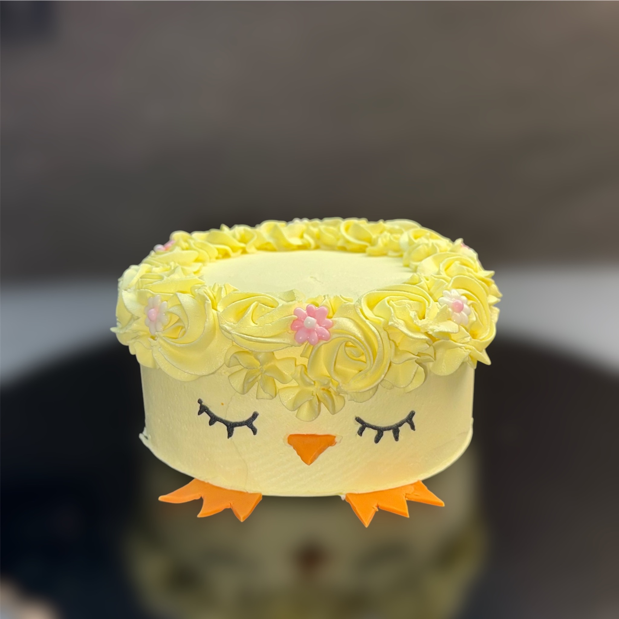 Uroczy tort w kształcie kurczaczka z żółtą kremową dekoracją, czarnymi rzęsami i pomarańczowym dziobem, na ciemnym tle.