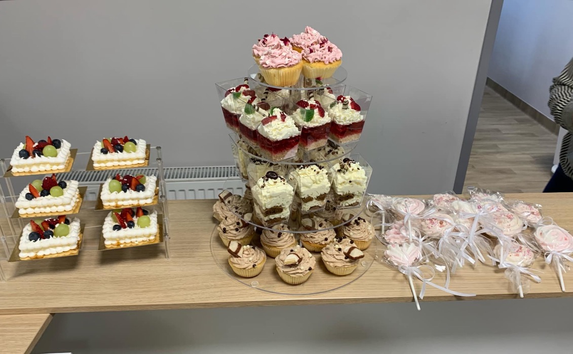 Słodki stół z różnorodnymi deserami: babeczki z kremem, mini torciki z owocami, desery w kubeczkach i lizaki bez dekoracji na jasnym blacie.