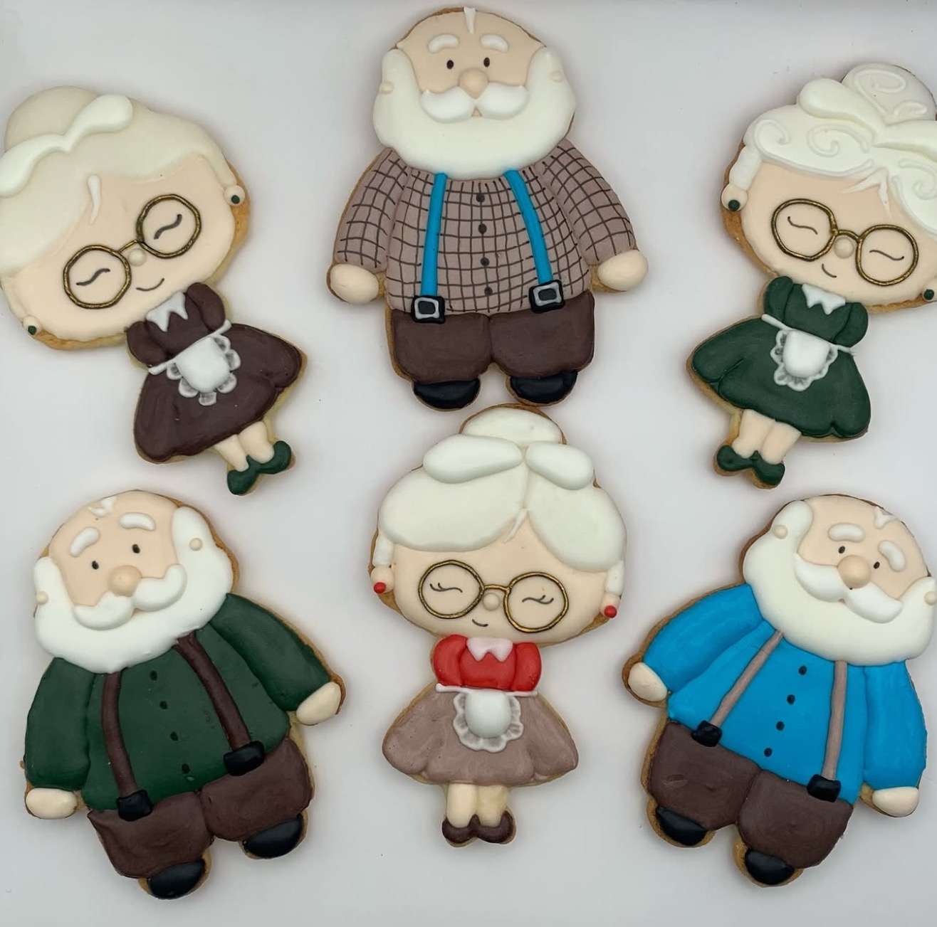 Ręcznie dekorowane pierniczki w kształcie uroczych staruszków, każdy z unikalnymi detalami stroju i wyrazem twarzy, ułożone na białym tle.
