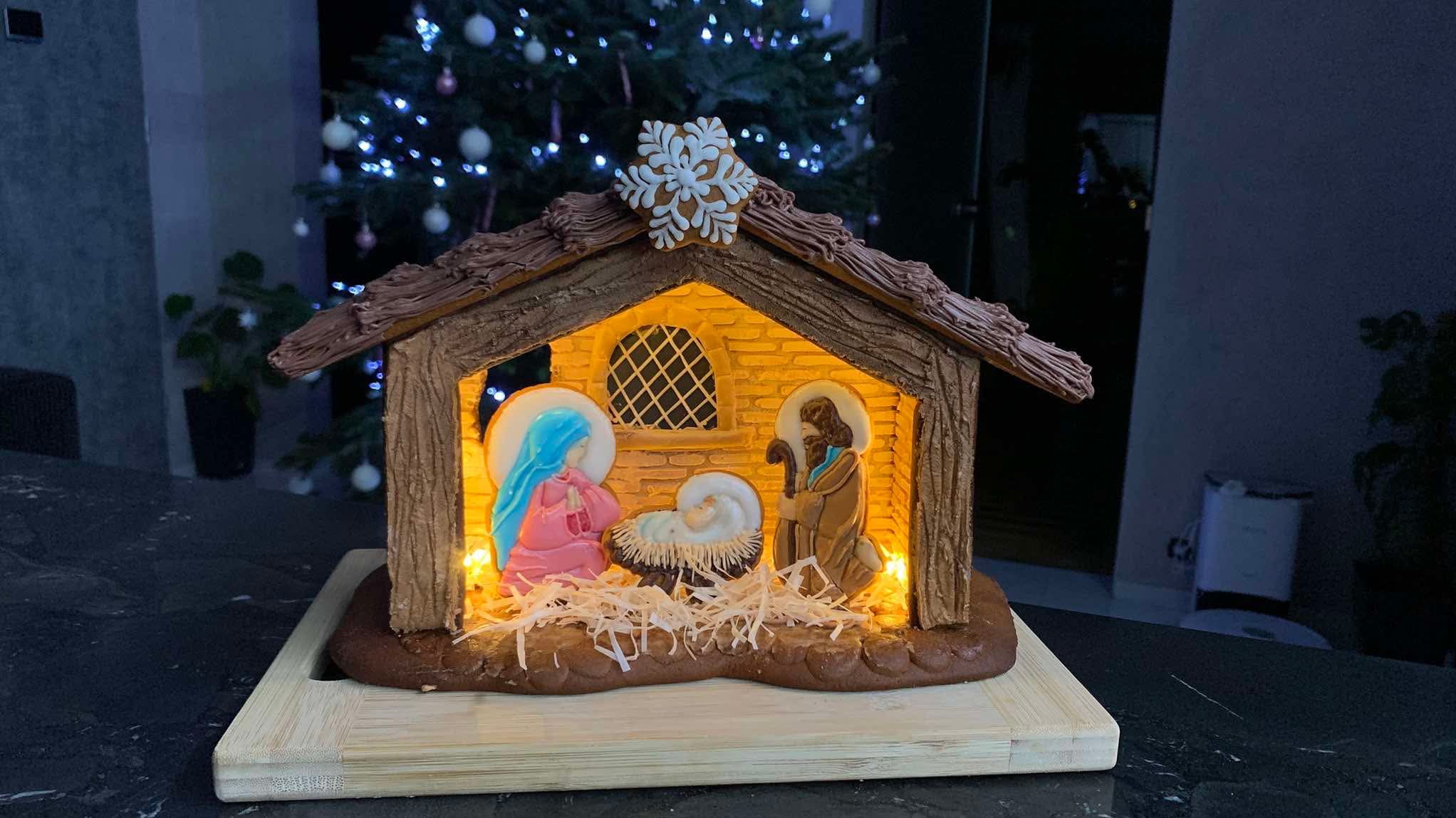 Piernikowa szopka bożonarodzeniowa z podświetleniem LED, na drewnianej desce, z choinką w tle. Dekoracja świąteczna z motywem religijnym, ręcznie robiona.