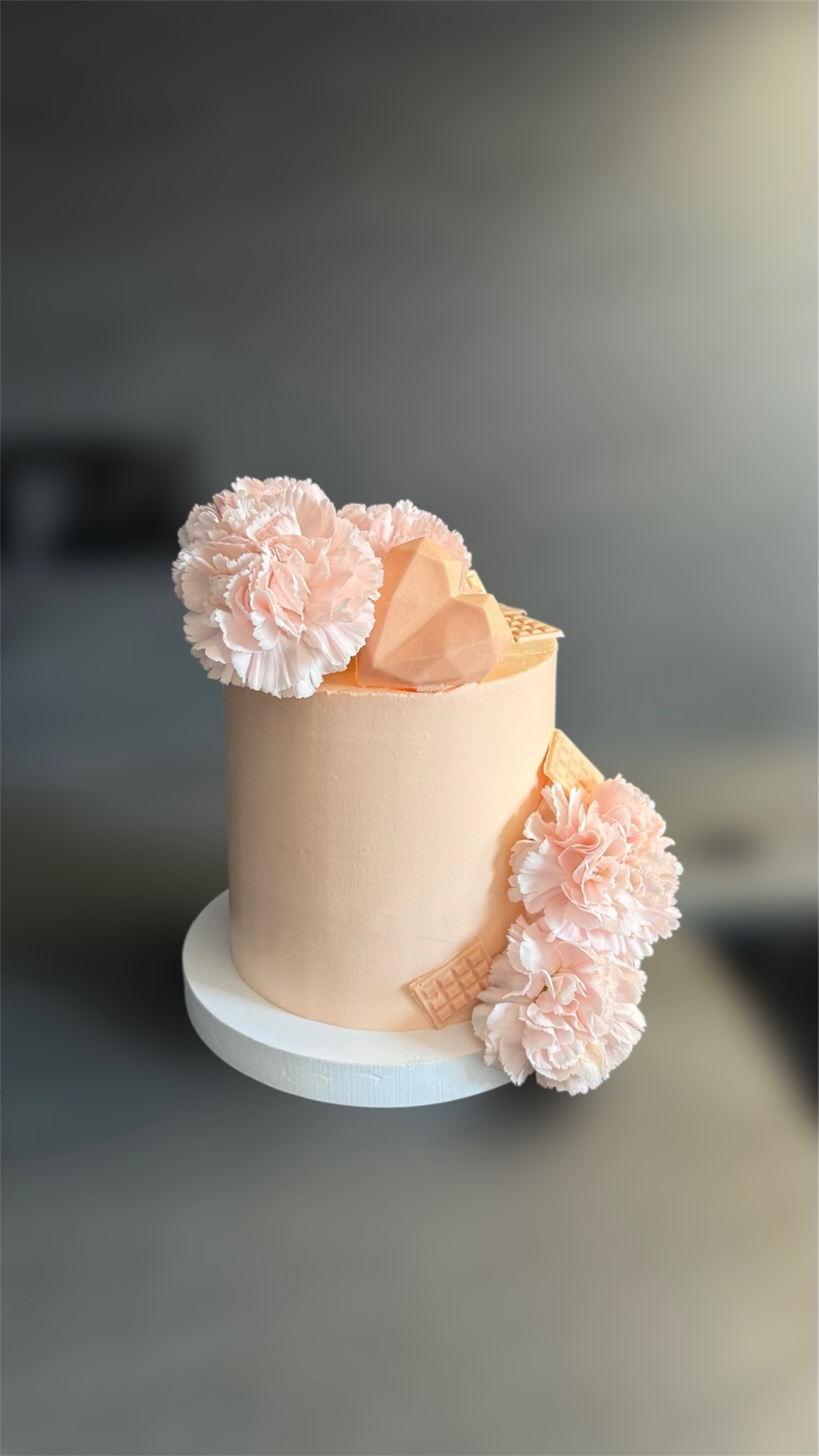 Delikatny tort w odcieniach brzoskwini, zdobiony jasnoróżowymi kwiatami i czekoladowym sercem, na białym podeście. Gładka powierzchnia i subtelne dekoracje.